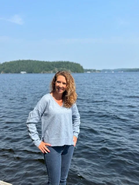 A woman with curly hair smiling and standing by a body of water, with a tree-covered island in the background on a sunny day.