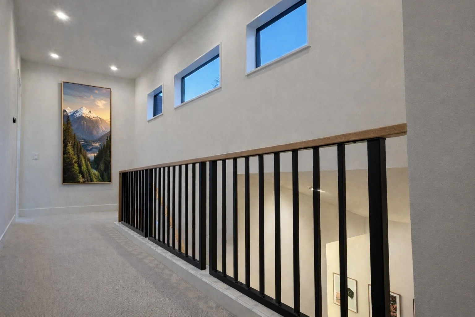 Interior of a modern home hallway with beige carpeting, white walls, a large landscape painting of mountains on the left wall, three small rectangular windows on the right wall, and a staircase with a black metal railing and wooden handrail leading d