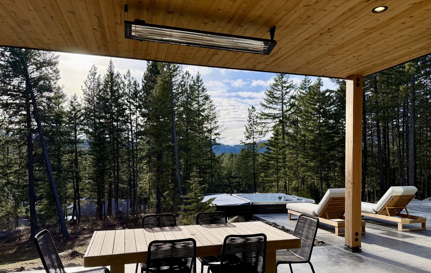 A covered outdoor patio with a wooden ceiling, a dining table with black chairs, two lounge chairs, a hot tub, and a forest of tall pine trees in the background under a partly cloudy sky.