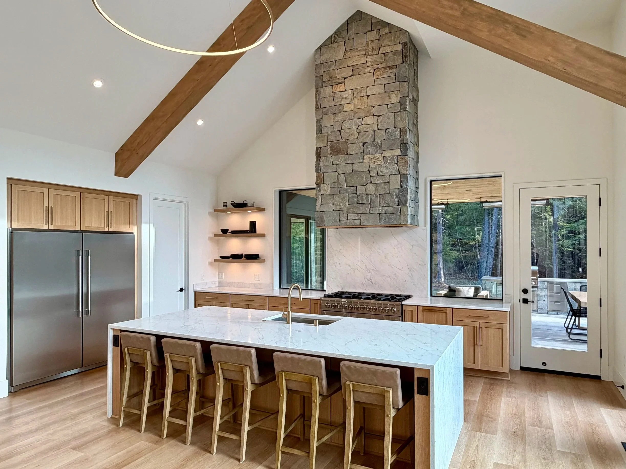 Modern kitchen with a large island, six chairs, wooden cabinets, stainless steel refrigerator, stone fireplace, large windows, and a door leading to an outdoor deck surrounded by trees.