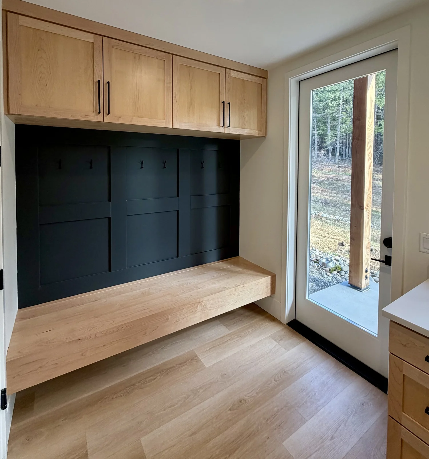 Interior of a room with a black wall with hooks, upper and lower wooden storage cabinets, a wooden bench, and a glass door leading outside to a natural landscape.