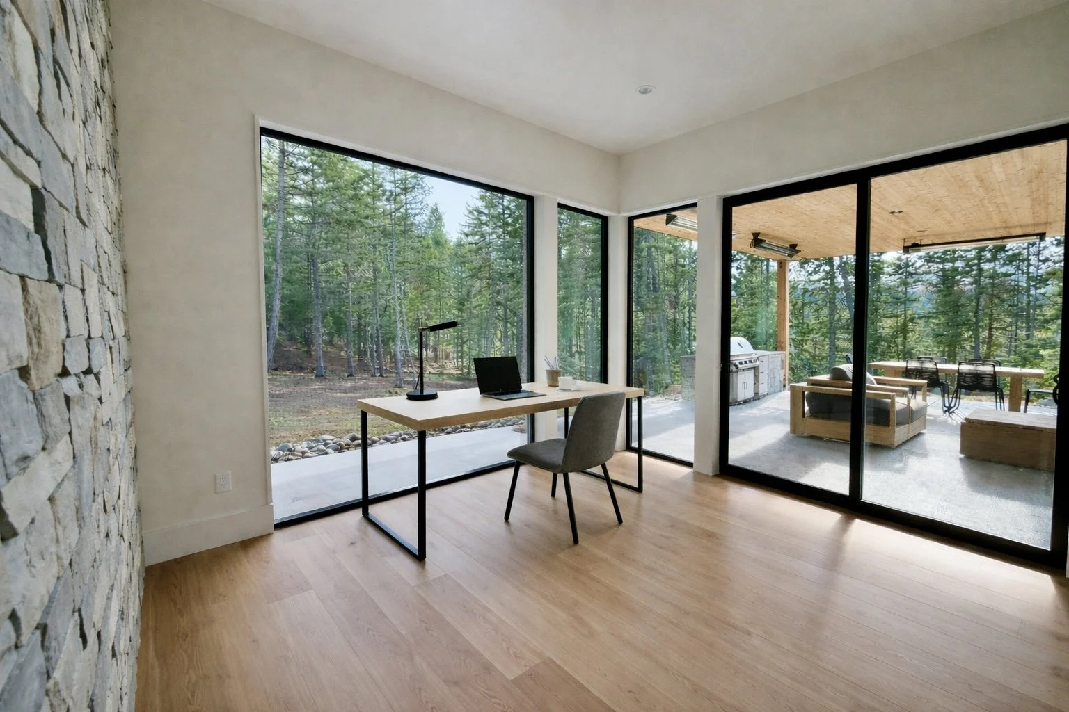A modern home office with a wooden desk, a laptop, a desk lamp, and a chair facing large glass windows overlooking a wooded outdoor patio space with outdoor furniture and a grill.