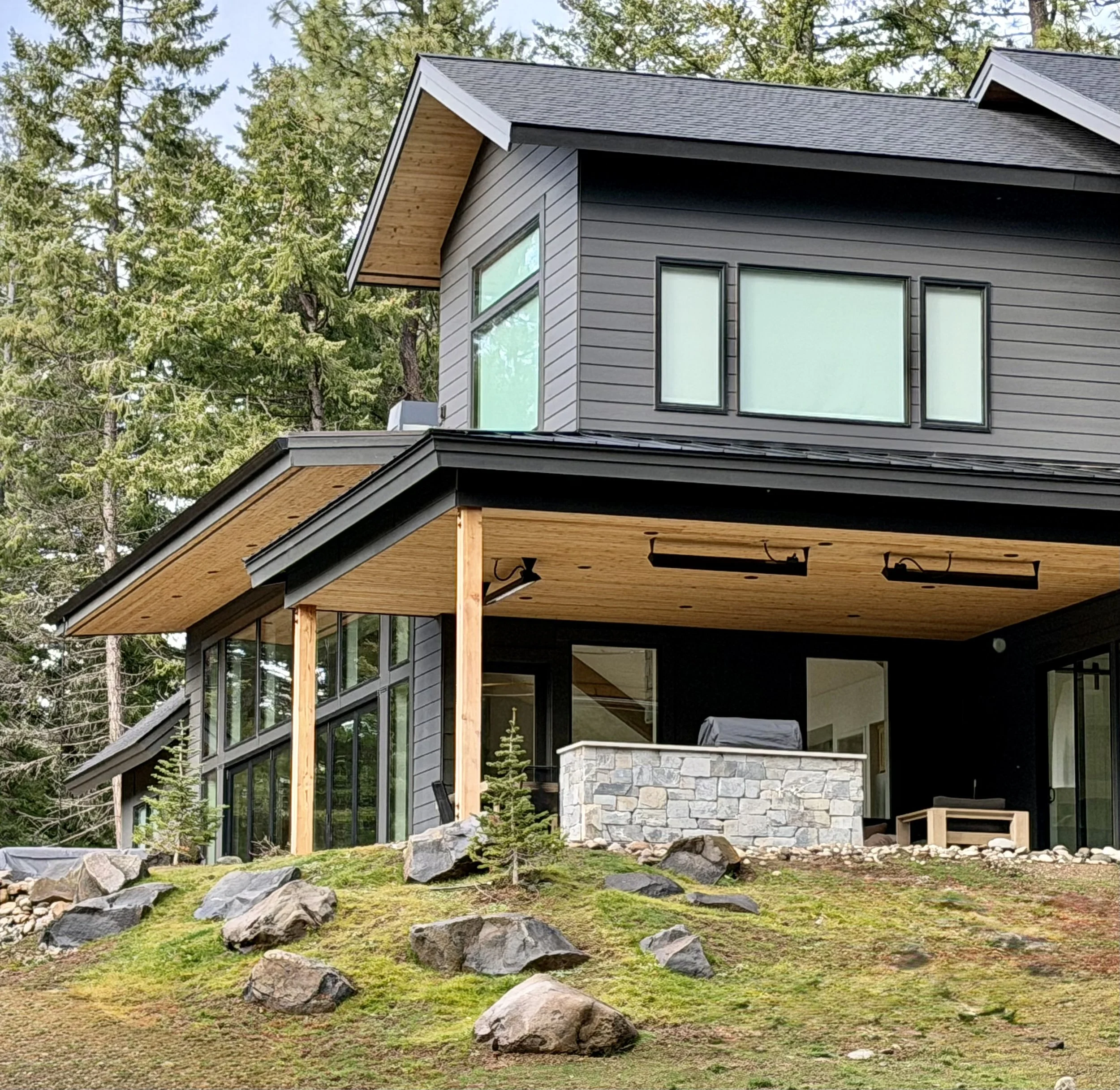 Modern two-story house with dark gray siding, large windows, and a covered patio with stone and wood accents, surrounded by trees and rocks on a hill.