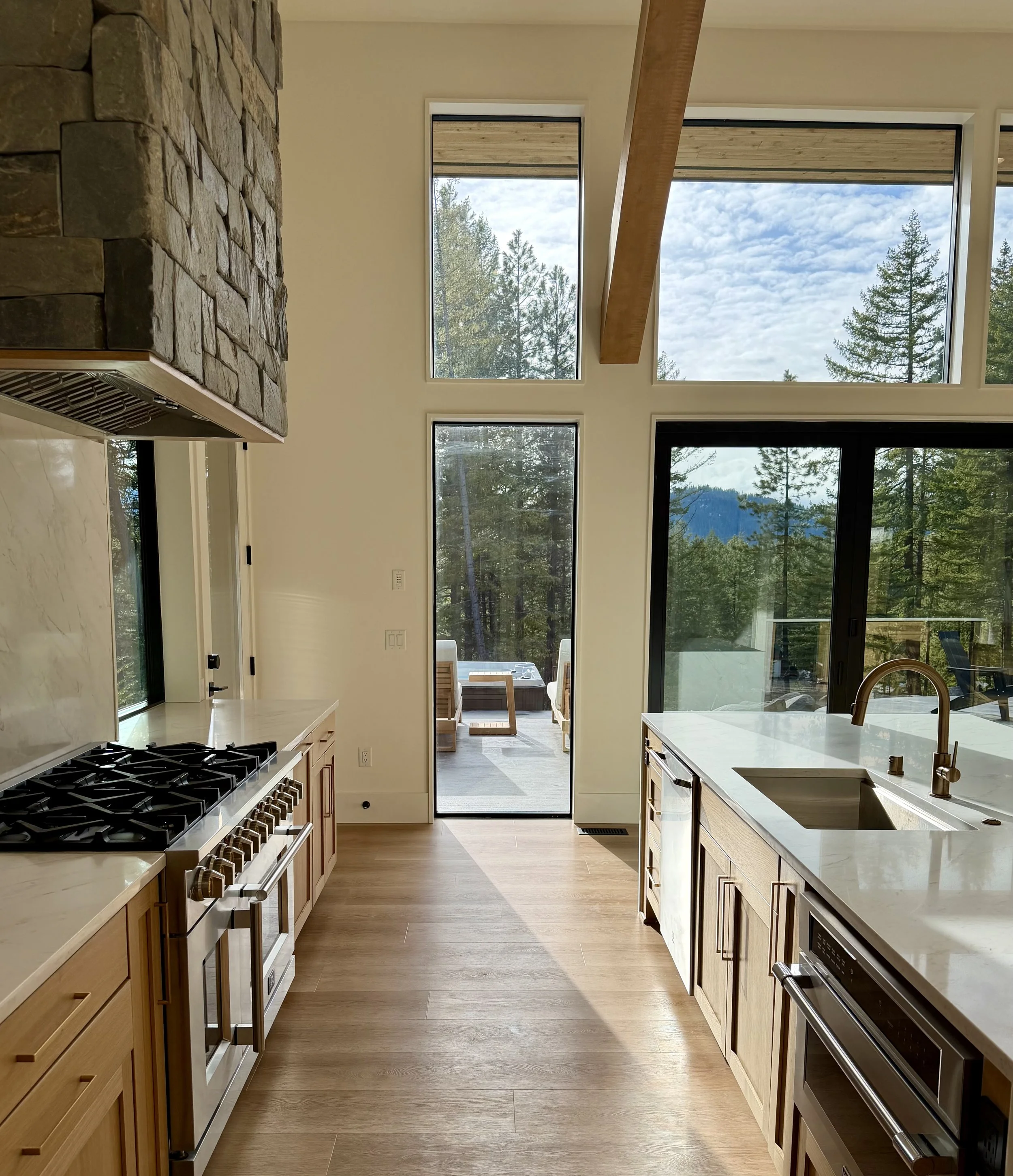 Modern kitchen with large windows and an outside patio with mountain views, featuring a gas stove, wooden cabinets, and a marble counter with a sink.