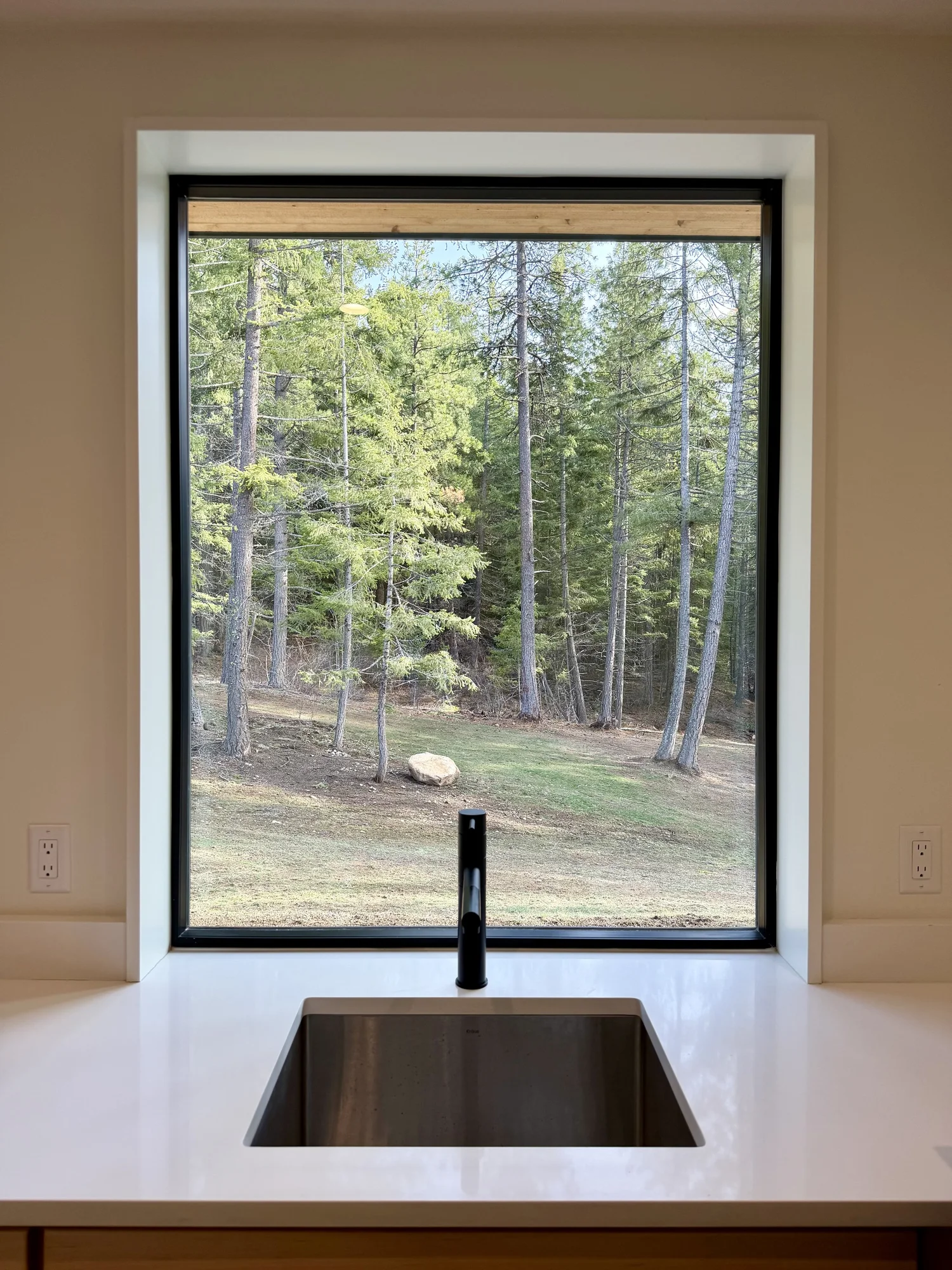 A kitchen view through a large window showing a wooded outdoor scene with trees, grass, and a large rock. The kitchen has white countertops, a stainless steel sink, and a black faucet.