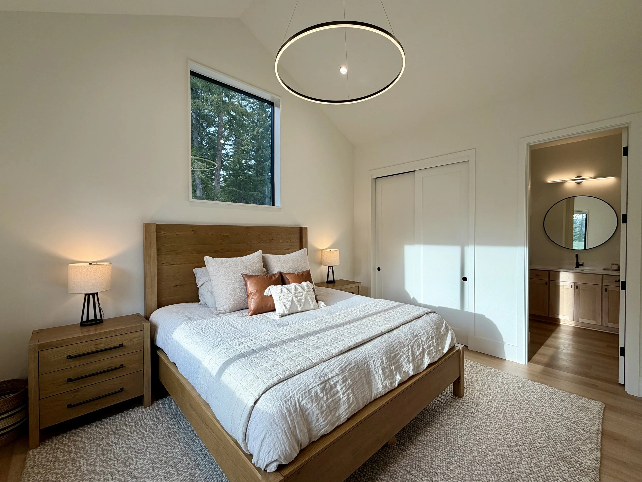 A bedroom with a wooden bed frame, white bedding, and multiple pillows. Two nightstands with lamps are on each side of the bed. A large window above the bed shows a view of trees. A closet with sliding doors is next to the bathroom, which has a sink 