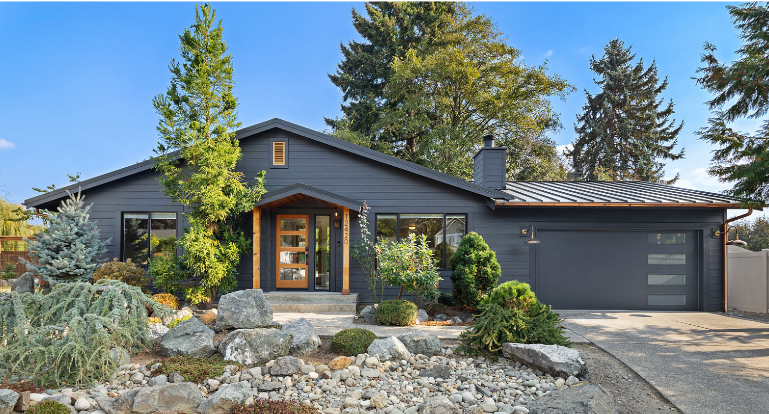 A modern, dark gray house with a landscaped front yard featuring rocks, shrubs, and trees, and a concrete driveway.