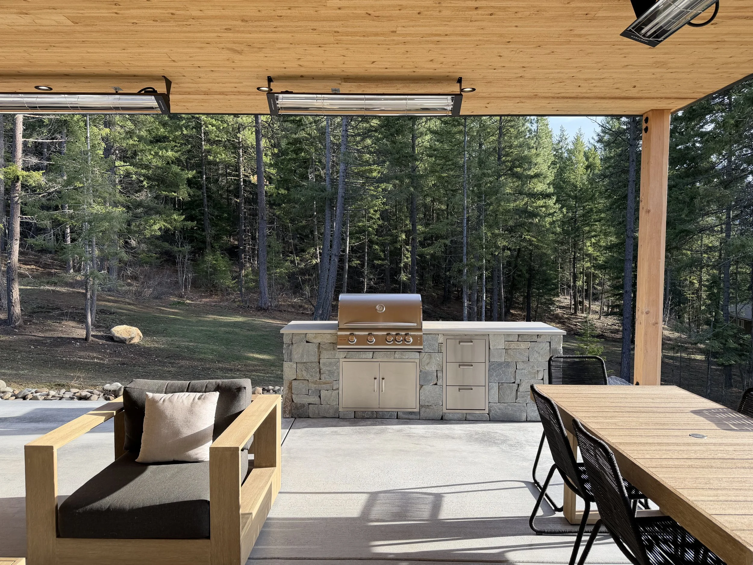 Outdoor patio with a stone barbecue grill, wooden dining table with black chairs, outdoor lounge chair with cushion, and a scenic forest background.