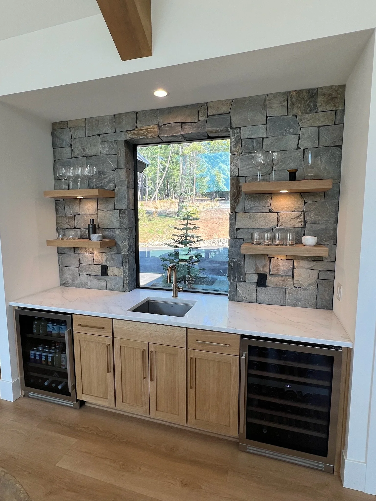 Kitchen bar area with a stone accent wall, window view of trees, wooden shelves with glassware, light wood cabinets, and a wine fridge.