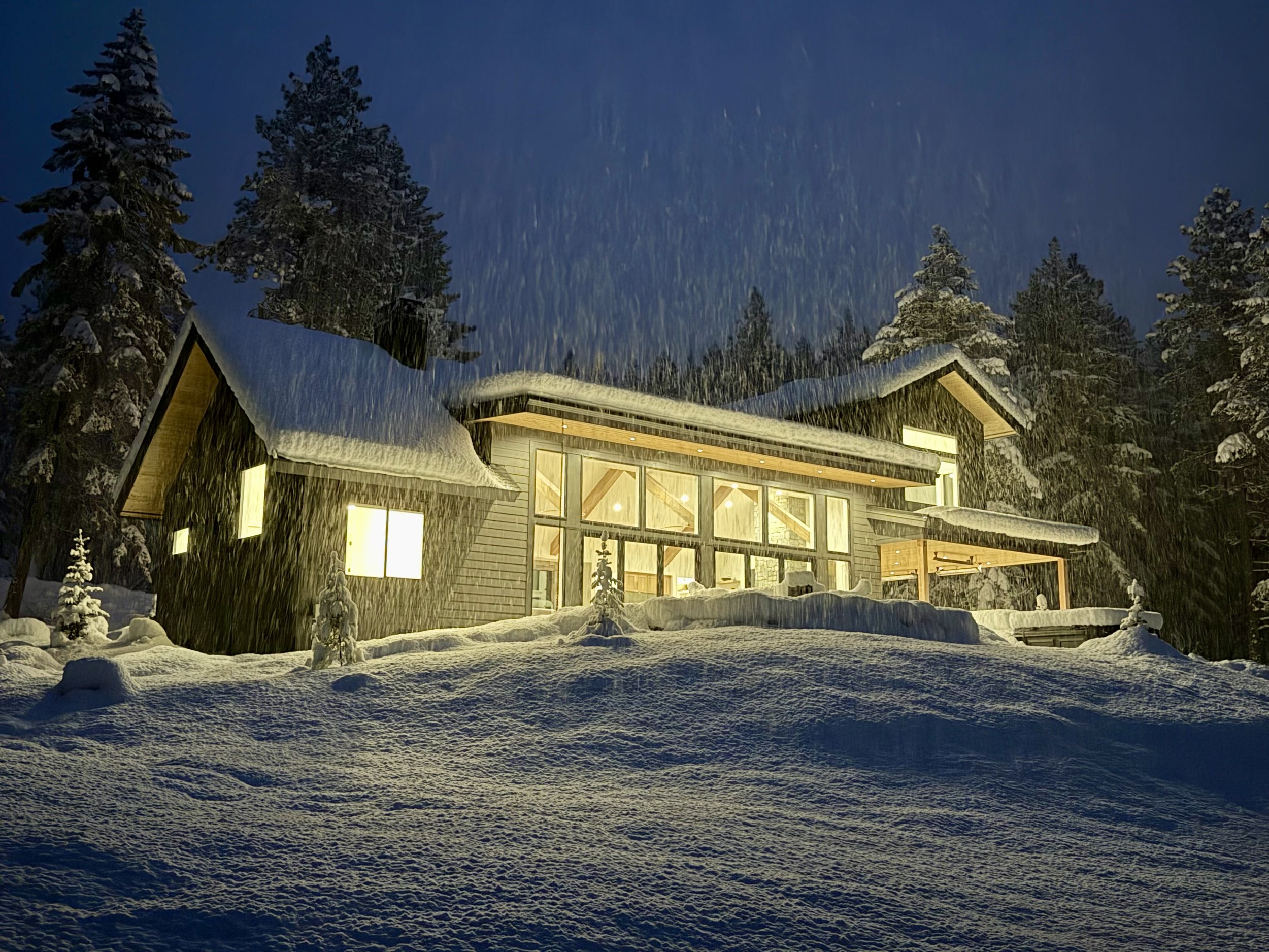A modern house illuminated from within, surrounded by snow and tall, snow-covered trees at night with snow falling.