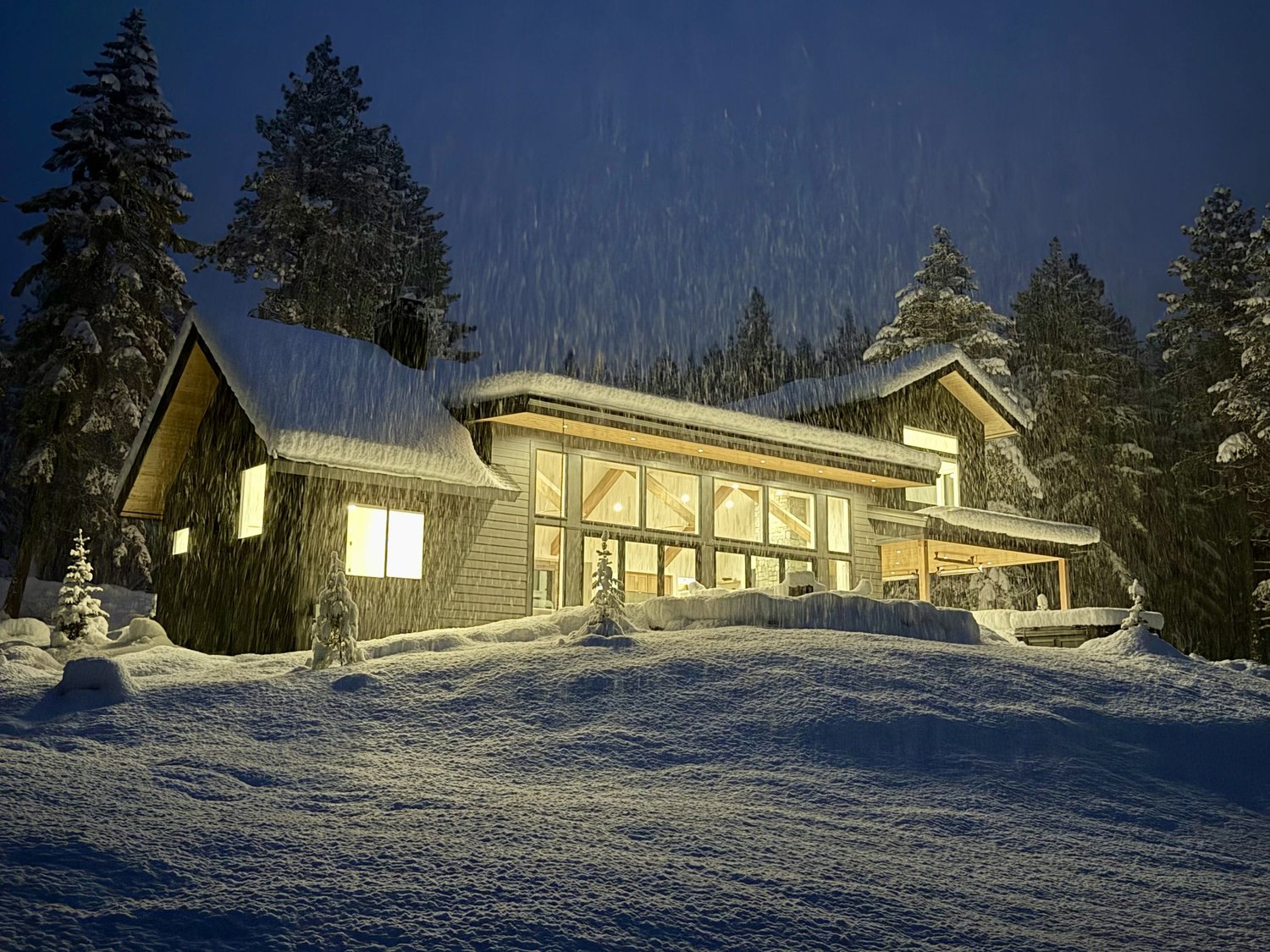 A modern house illuminated from within, surrounded by snow and tall, snow-covered trees at night with snow falling.