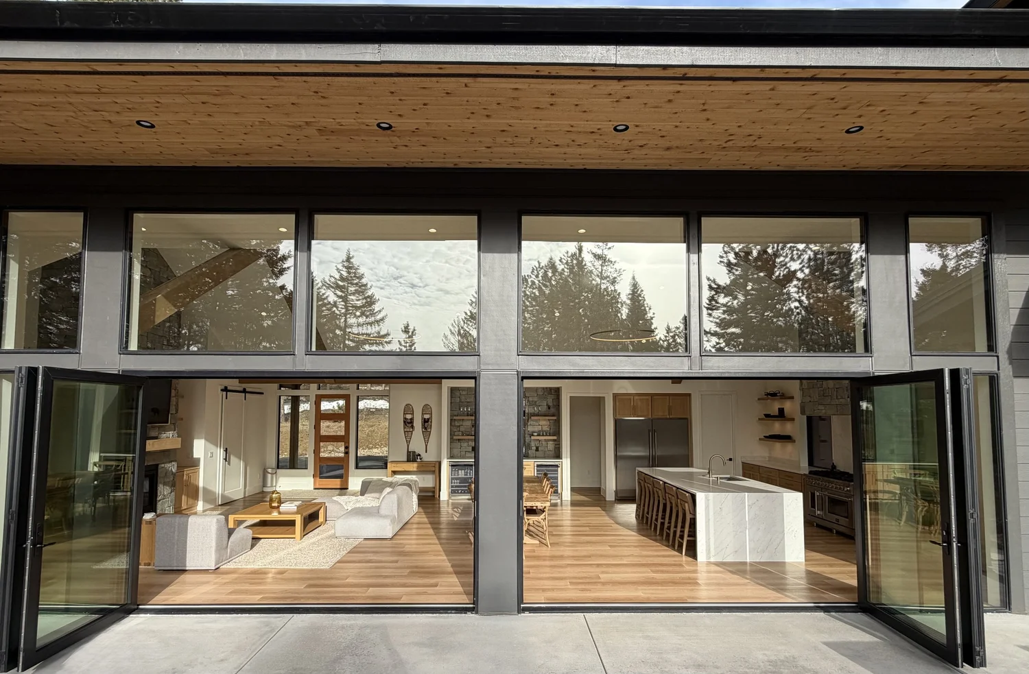Open sliding glass doors revealing a modern living room and kitchen inside a house with large windows and a wooden ceiling, surrounded by trees.