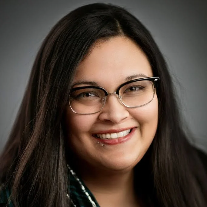 Close-up of a smiling woman with long dark hair, glasses, and a piercing on her lower lip, against a neutral background.