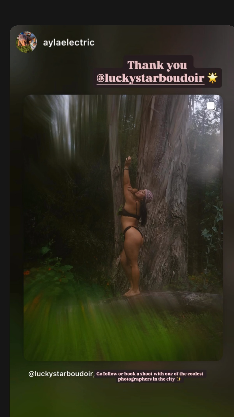 A woman in black underwear standing in front of a large tree outside, reaching upward with one arm, wearing a pink headscarf, in a forest setting.