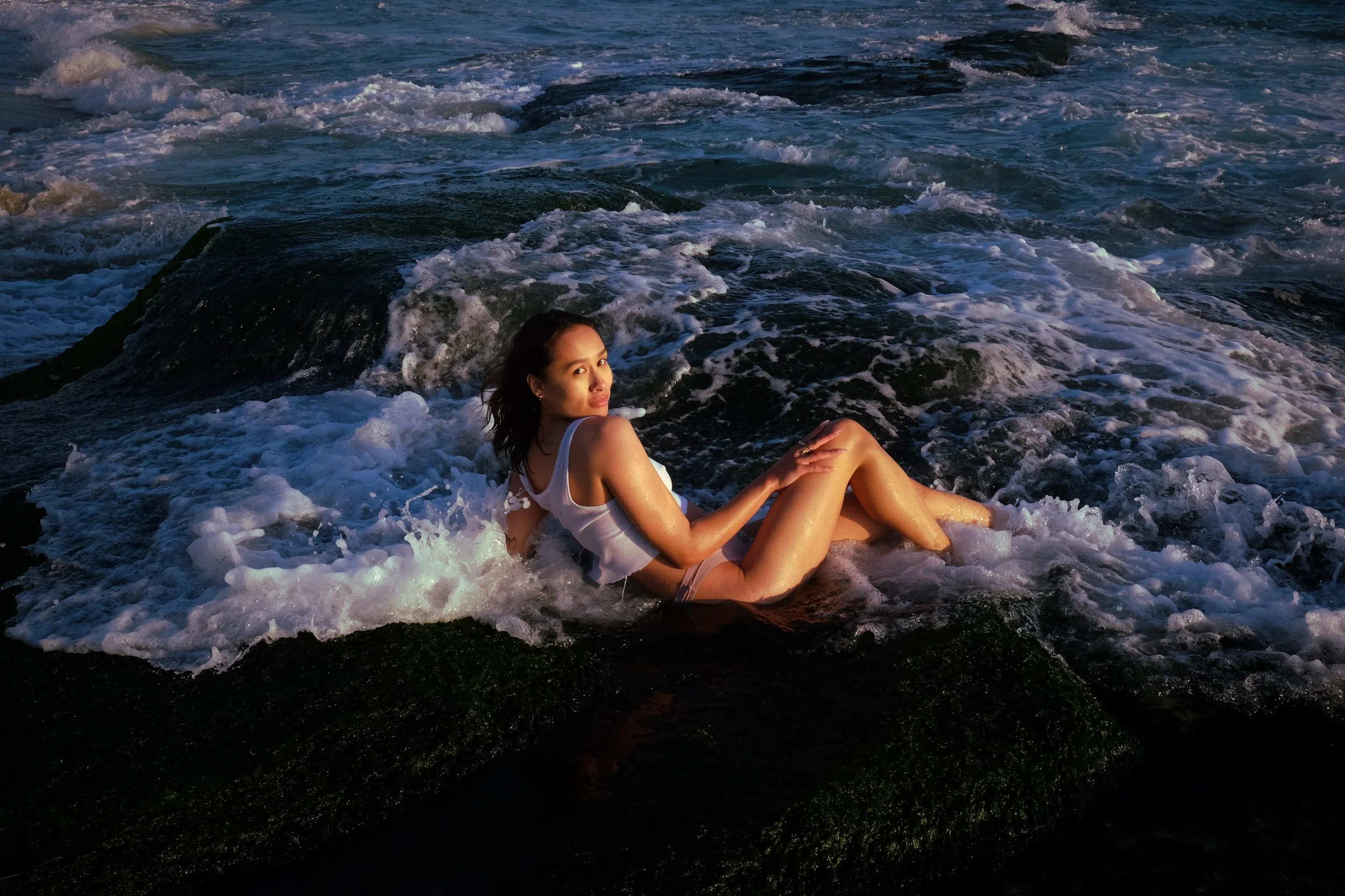 A woman in a white tank top and shorts sitting on rocks among foamy ocean waves on a beach at sunset.