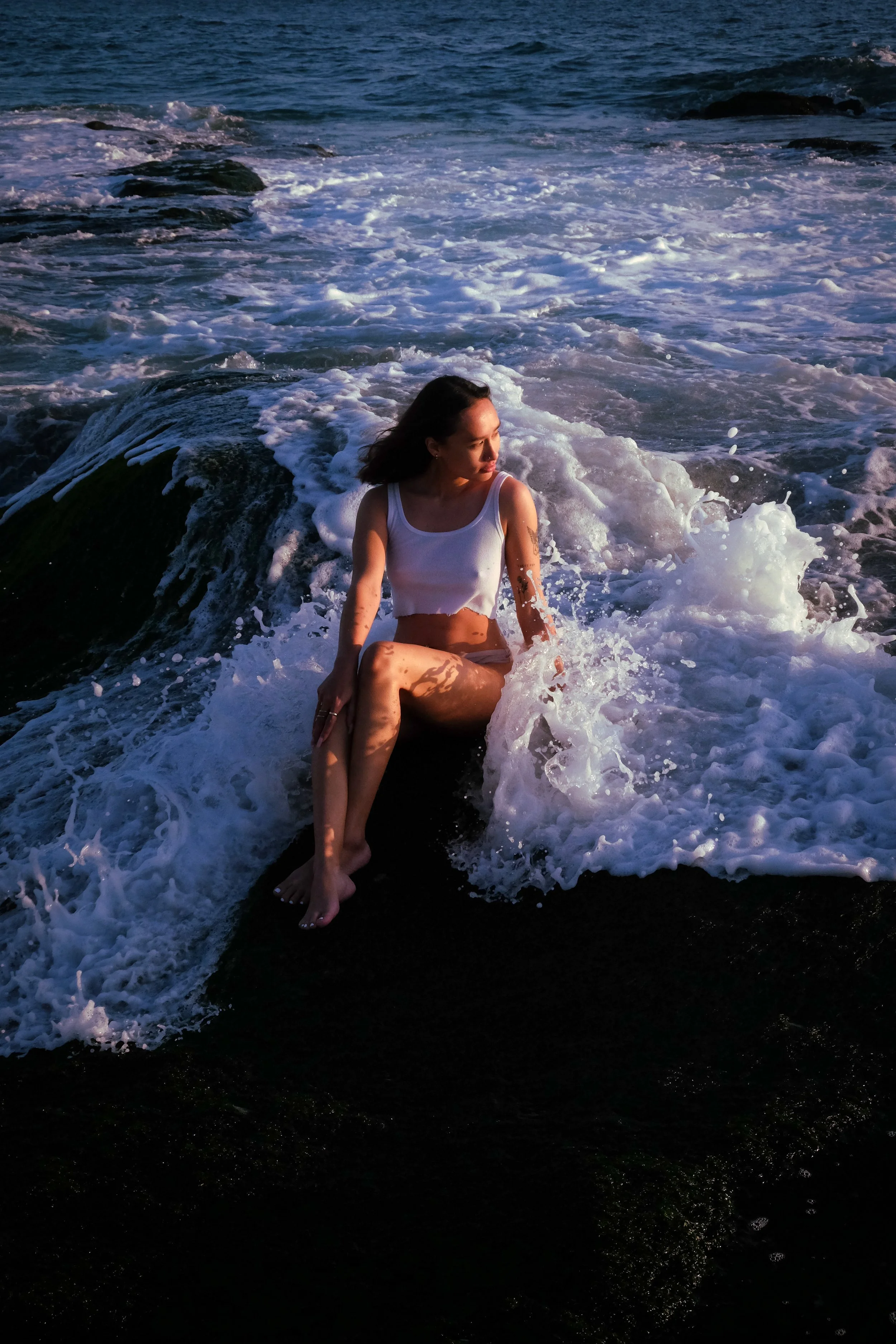 A woman in a white tank top sitting on a rock at the edge of the ocean with foamy waves surrounding her, during sunset or sunrise.