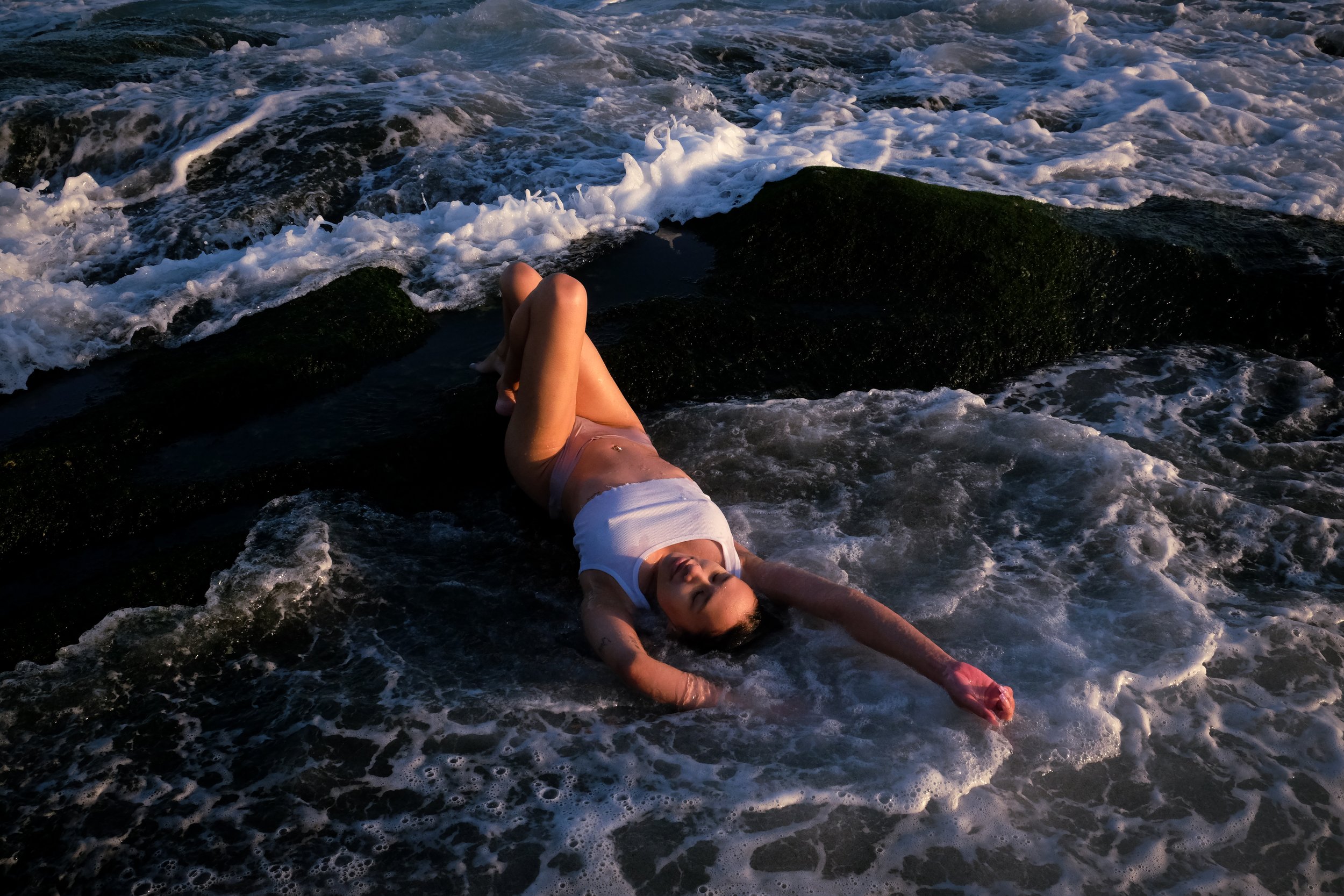 A woman lying on her back on rocks by the ocean, her eyes closed, with foamy waves around her during sunset.