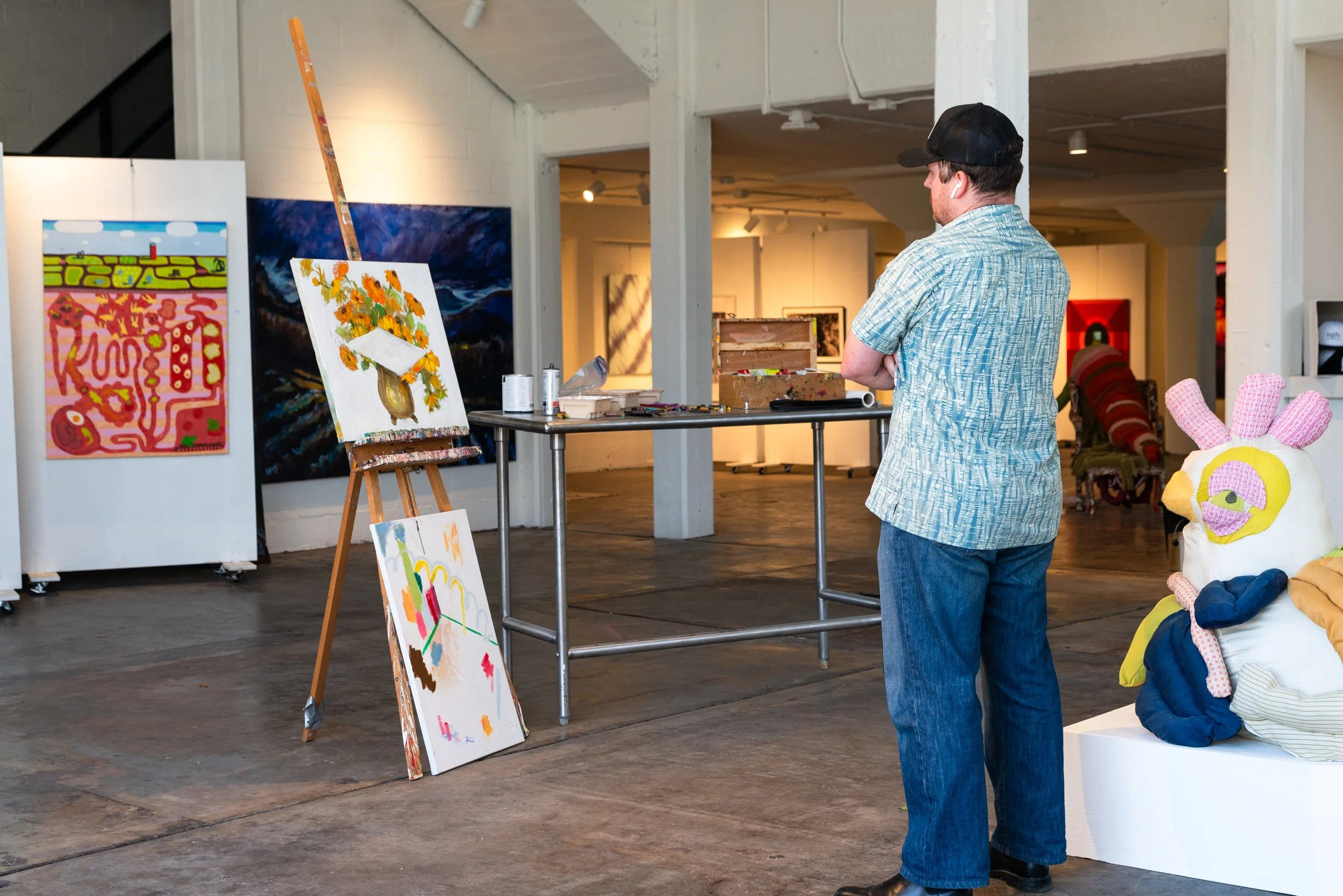 A man in a blue patterned shirt and a black cap standing in an art gallery with paintings on the walls, and colorful sculptures including a plush bunny and a face on a white platform.