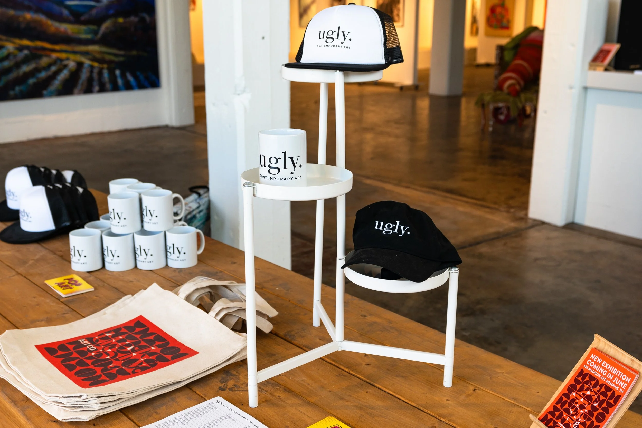 Display table with merchandise including white mugs, black and white caps, tote bags, and promotional signs at an art gallery.
