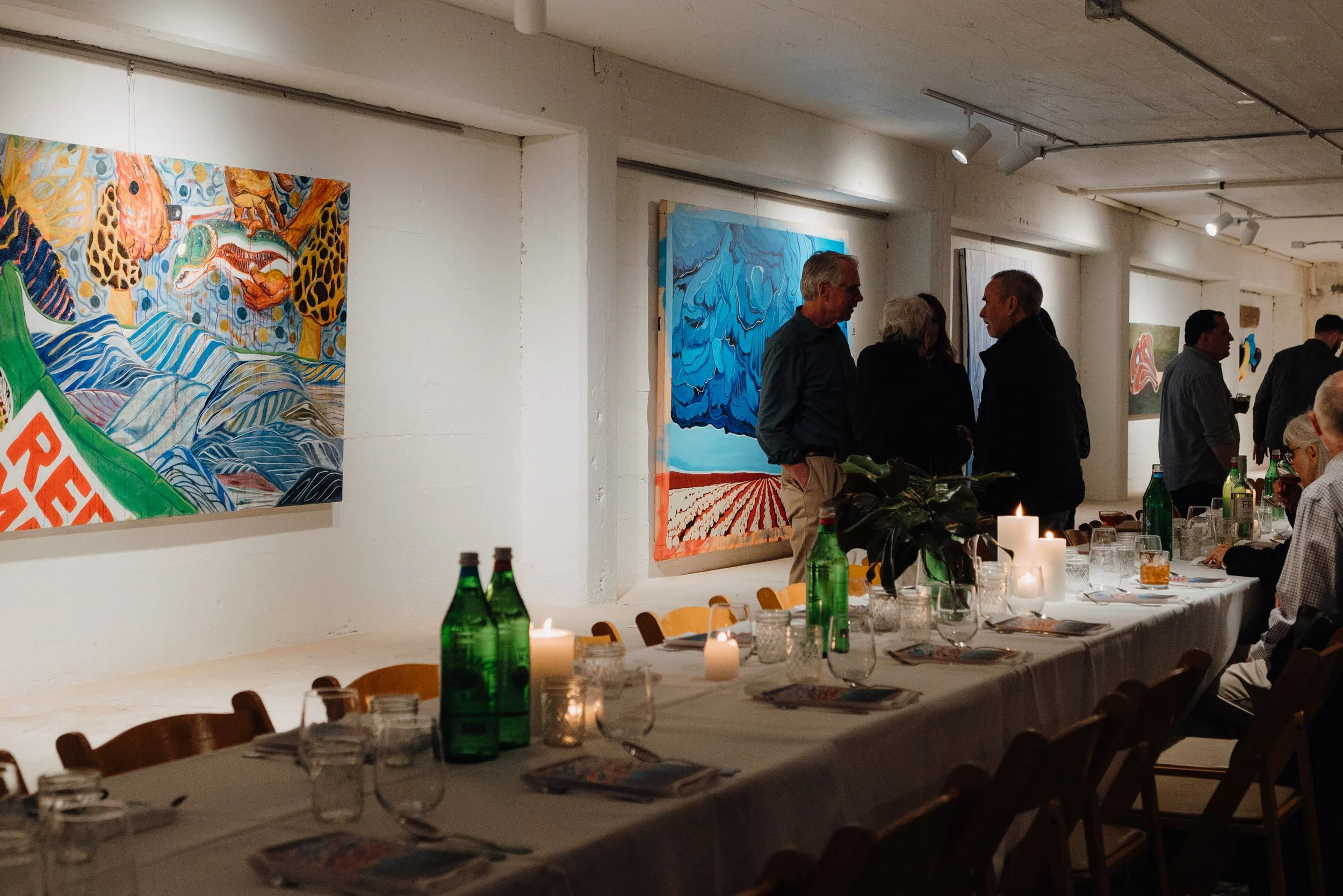 People socializing at an art gallery with vibrant paintings on the wall and a long dinner table with candles and drinks.