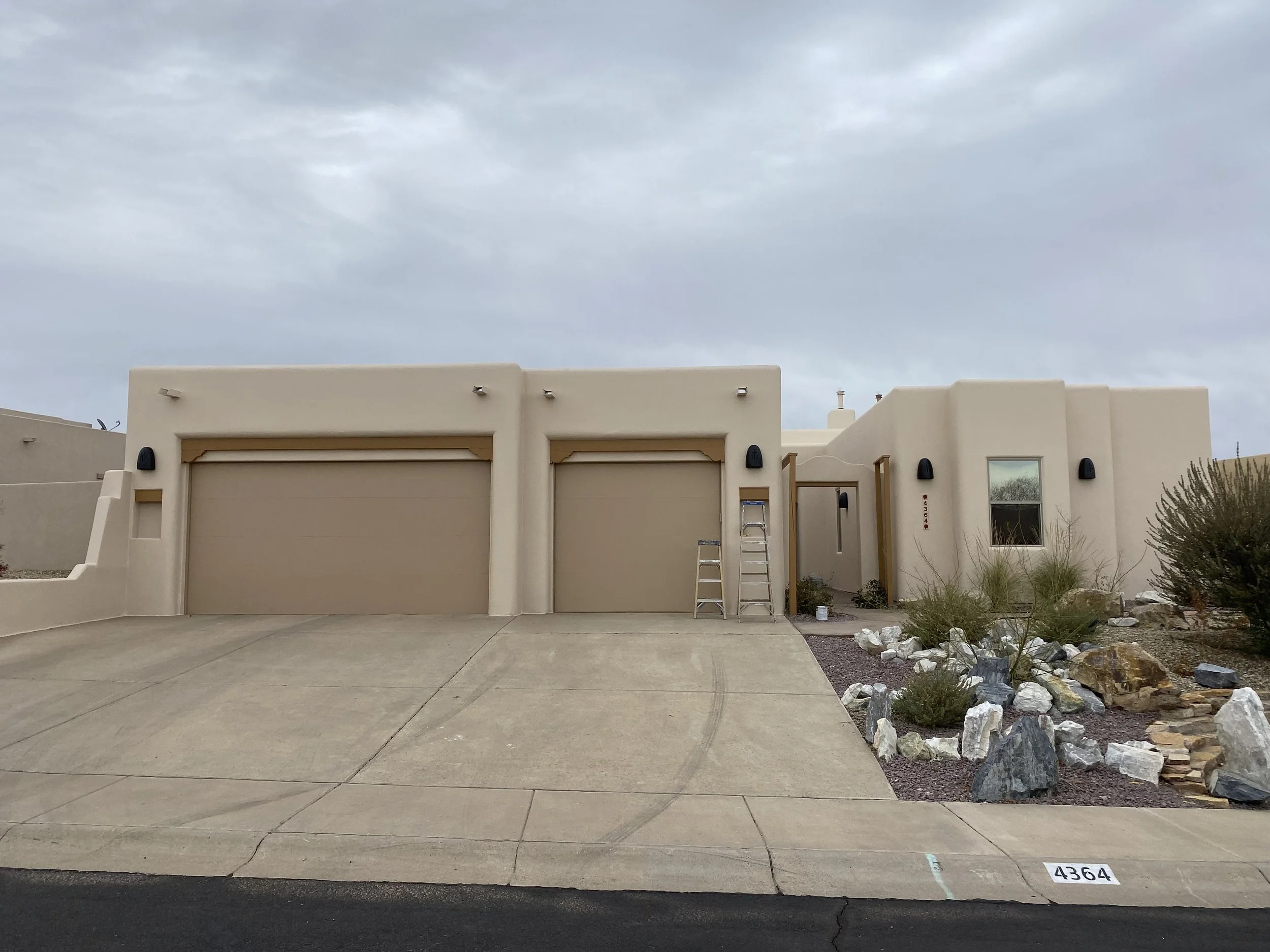 View of modern beige building with garage doors, ladder, and landscaping including rocks and desert plants, under cloudy sky. Fresh elastomeric stucco coating.
