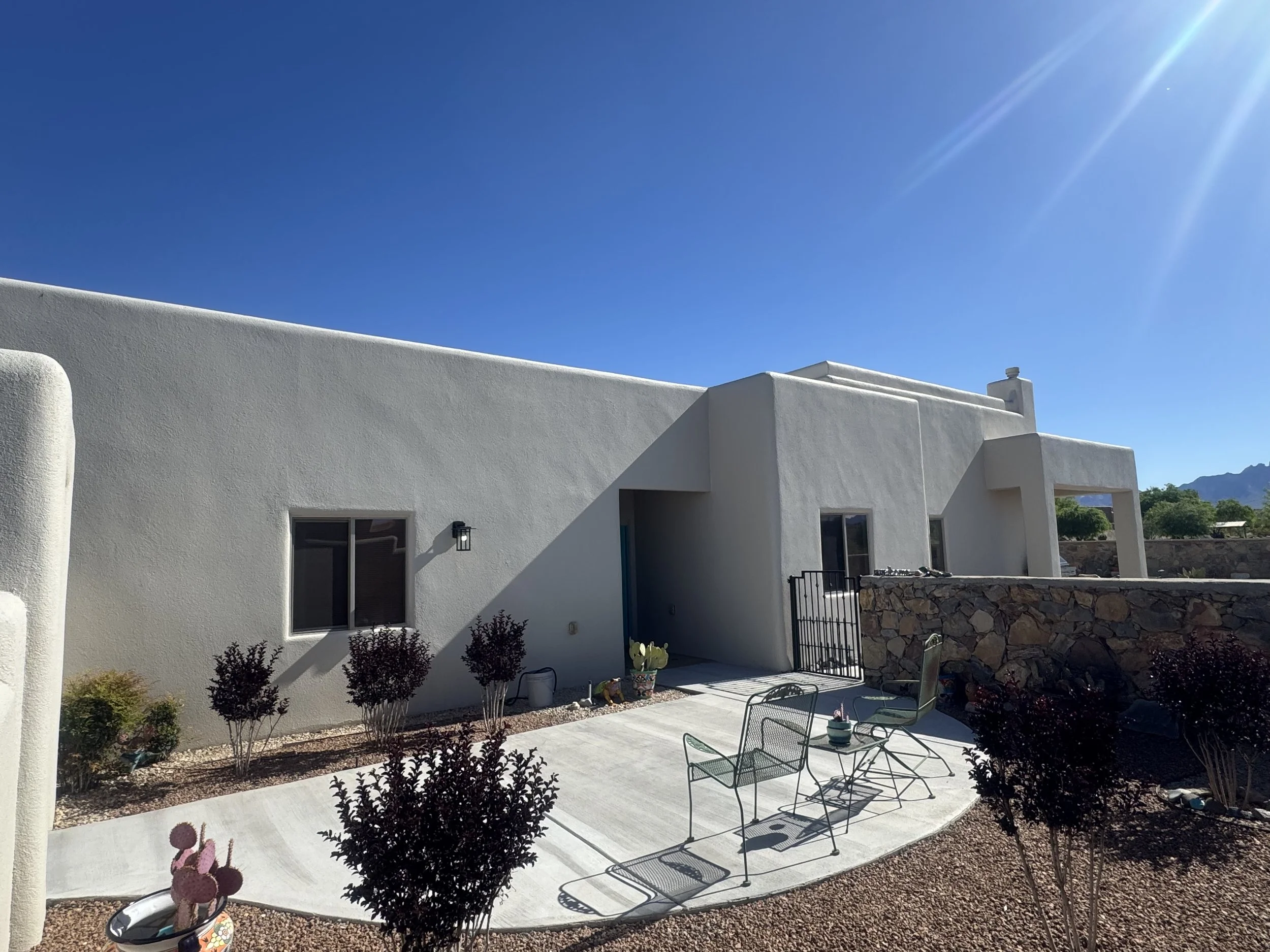 White stucco house with flat roof, black windows, and a black gate. Front yard with small plants, cactus, patio furniture, and a bright sunny sky with lens flare. Fresh elastomeric stucco coating.