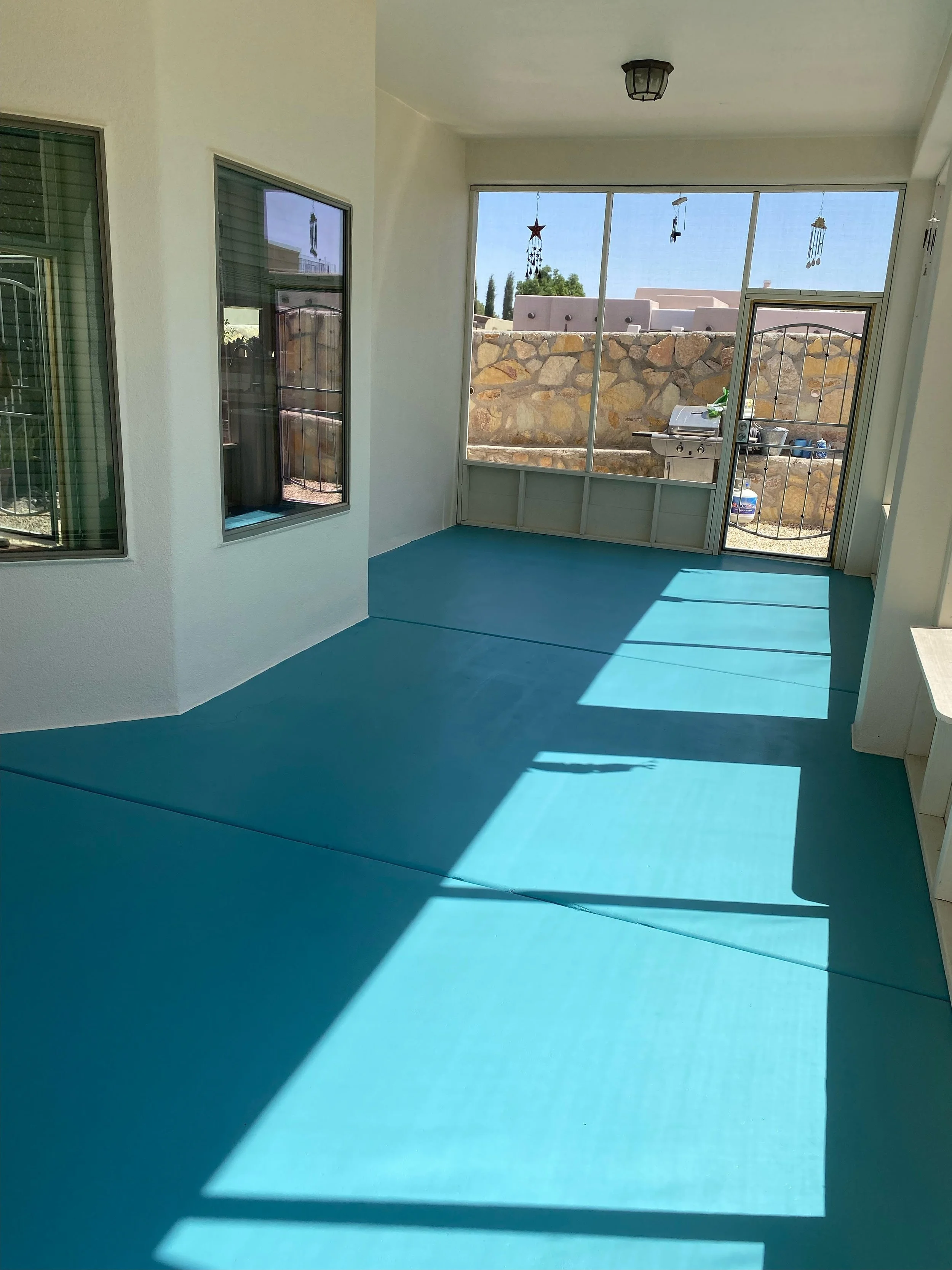 A sunlit balcony with a screened door, glass windows, and a view of a stone wall, patio, and outdoor grill. Custom concrete staining.