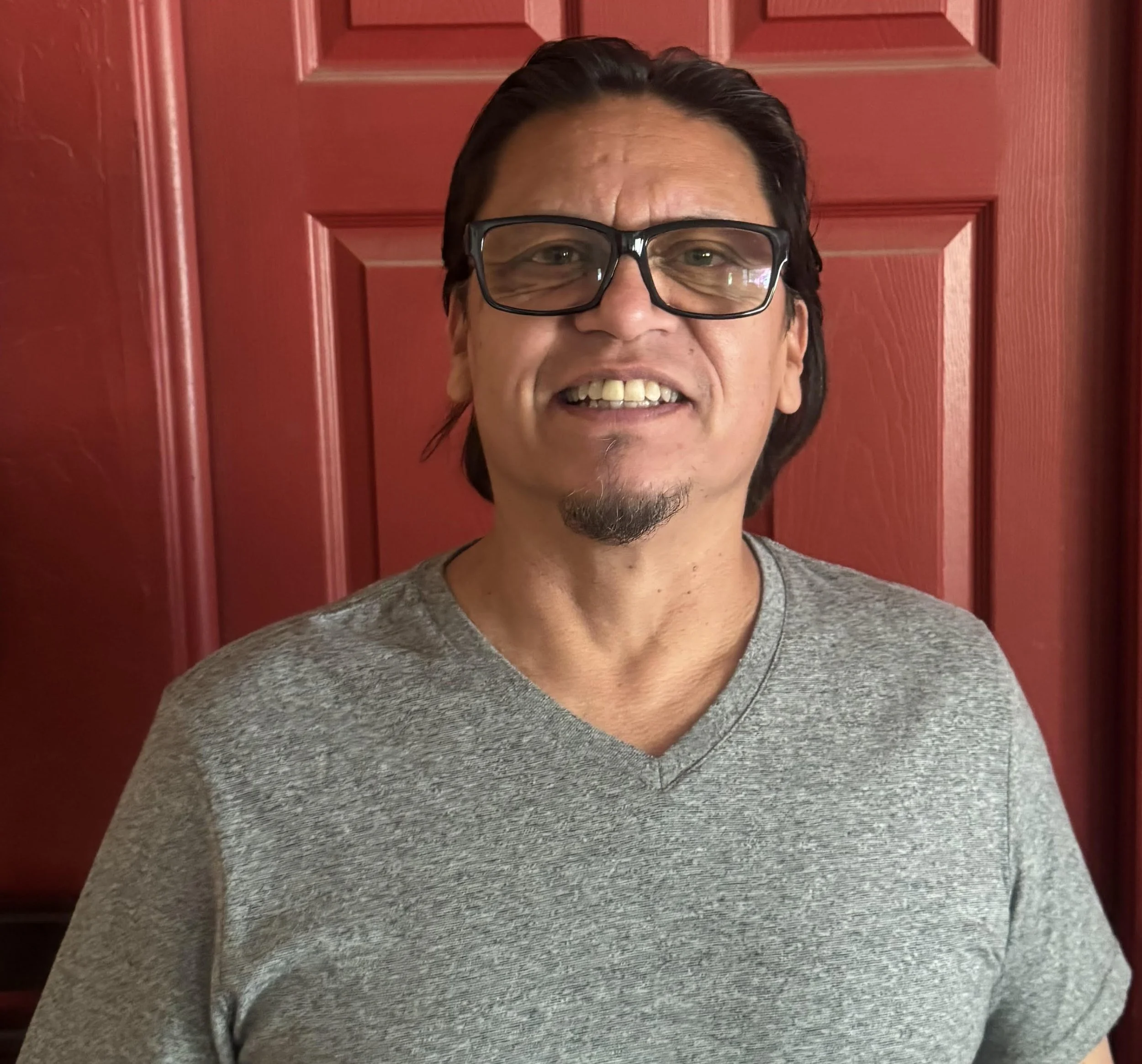 A smiling man with glasses, dark hair, and facial hair, standing in front of a red door, wearing a gray shirt.