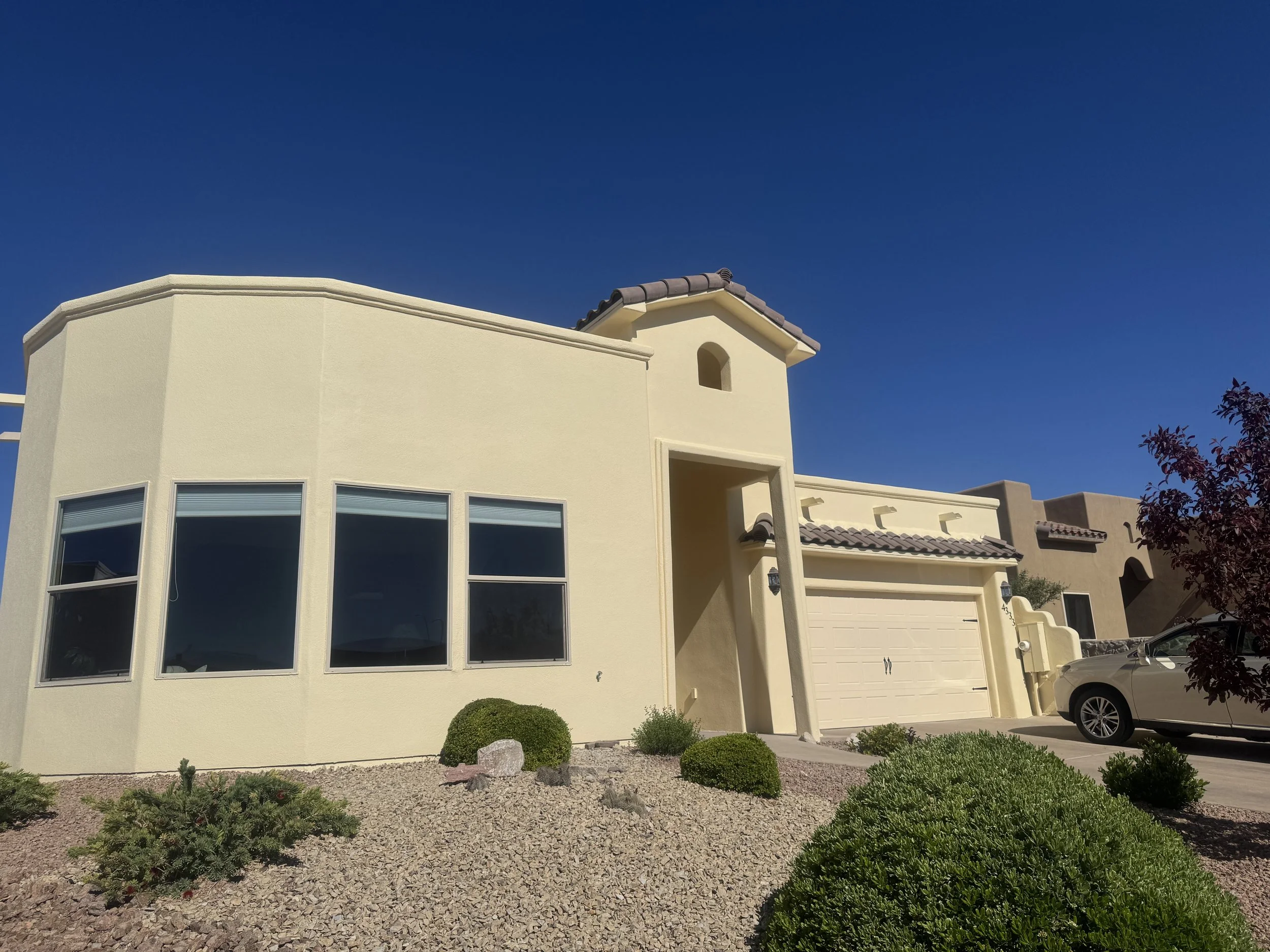 A modern cream-colored house with multiple large windows, a small tower-like structure with a small arched window, a garage door, and a driveway with parked cars. The front yard has minimal landscaping with small bushes, rocks, and gravel mulch. The sky is clear and blue.