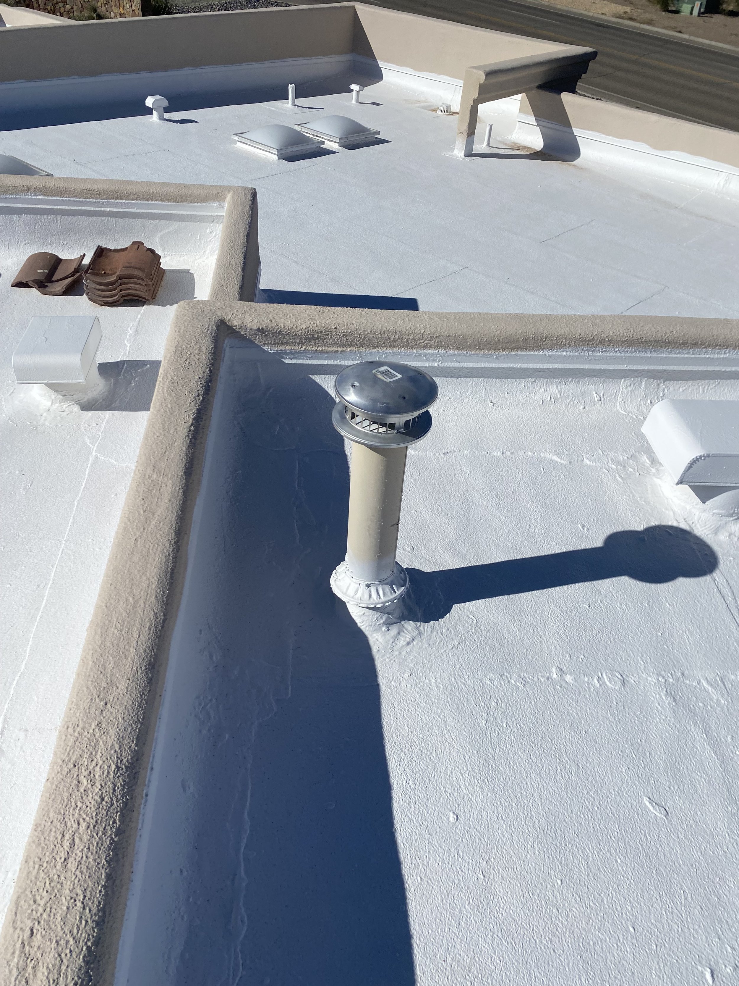 The roof has a parapet wall around the edge, and a shadow of a vertical pipe is cast on it. Part of a nearby road is visible in the background. Fresh elastomeric roof coating.