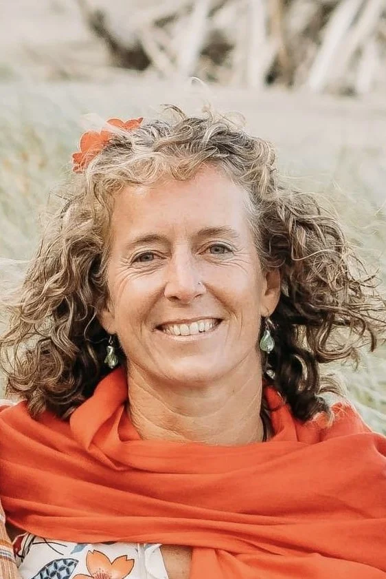A woman with curly hair smiling, wearing earrings and a red shawl, outdoors with a blurry natural background.