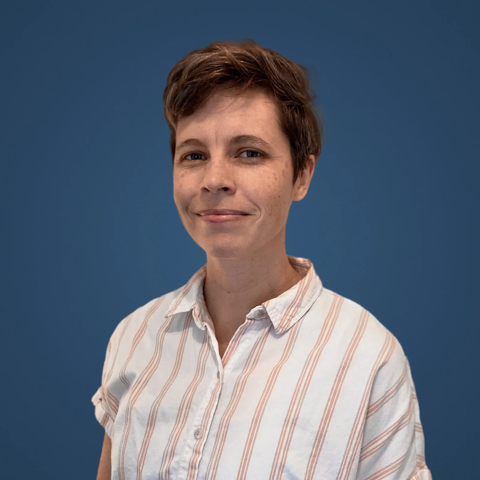 Woman with short brown hair wearing a white and beige striped shirt against a blue background.