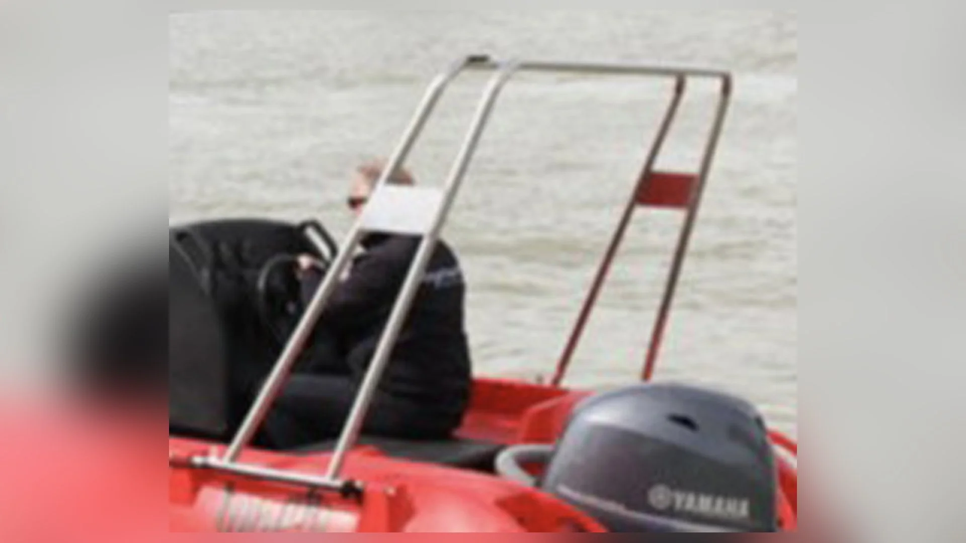 Person in a black jacket sitting on a red boat with a Yamaha outboard motor, near a body of water, with a metal frame structure on the boat.