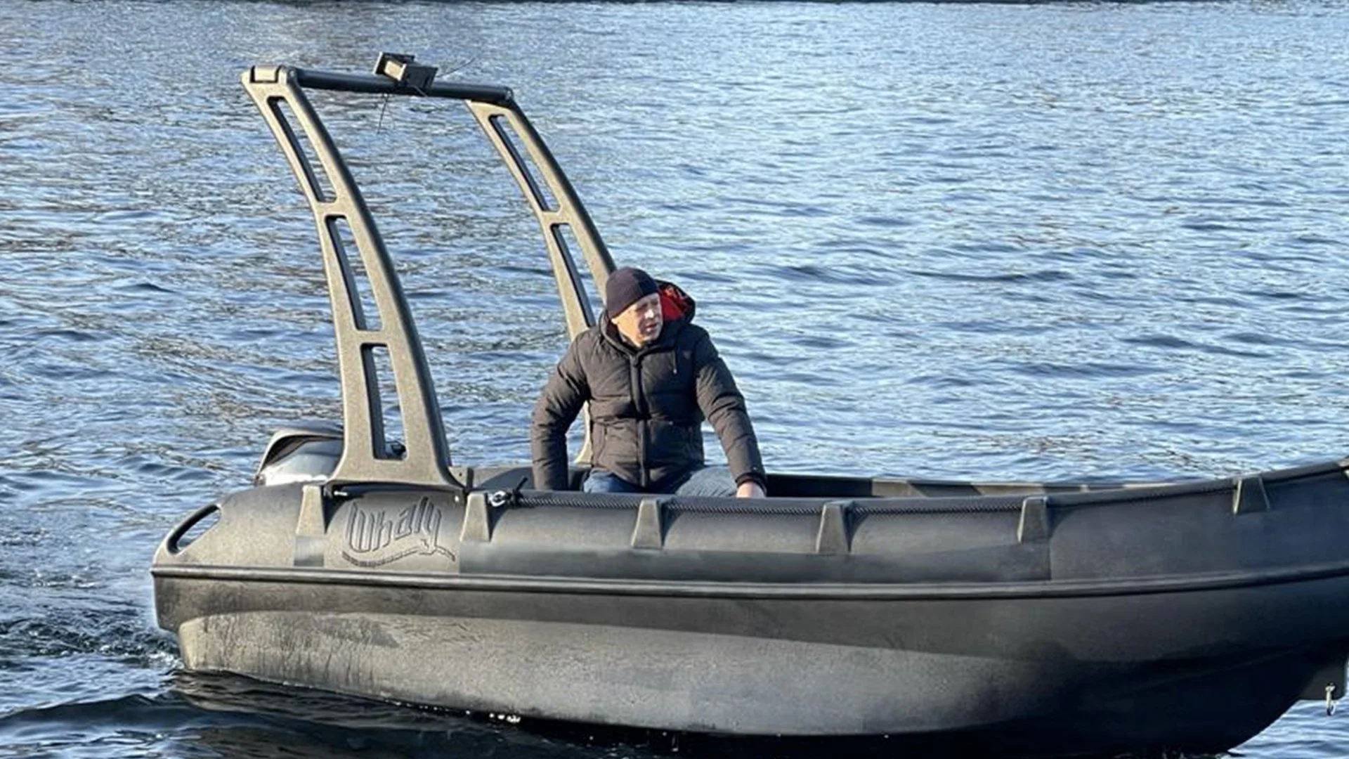 A man wearing a black jacket and a beanie sitting in a black inflatable boat on a body of water.