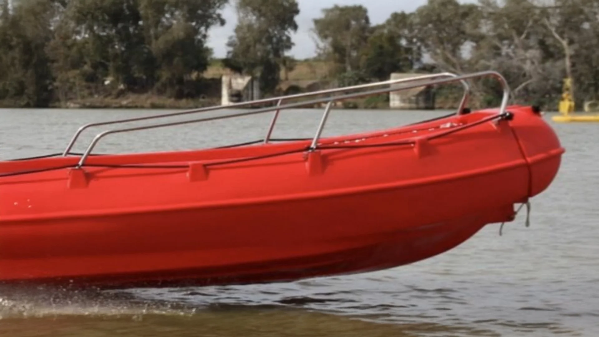Red boat on the water with trees and a bridge in the background.