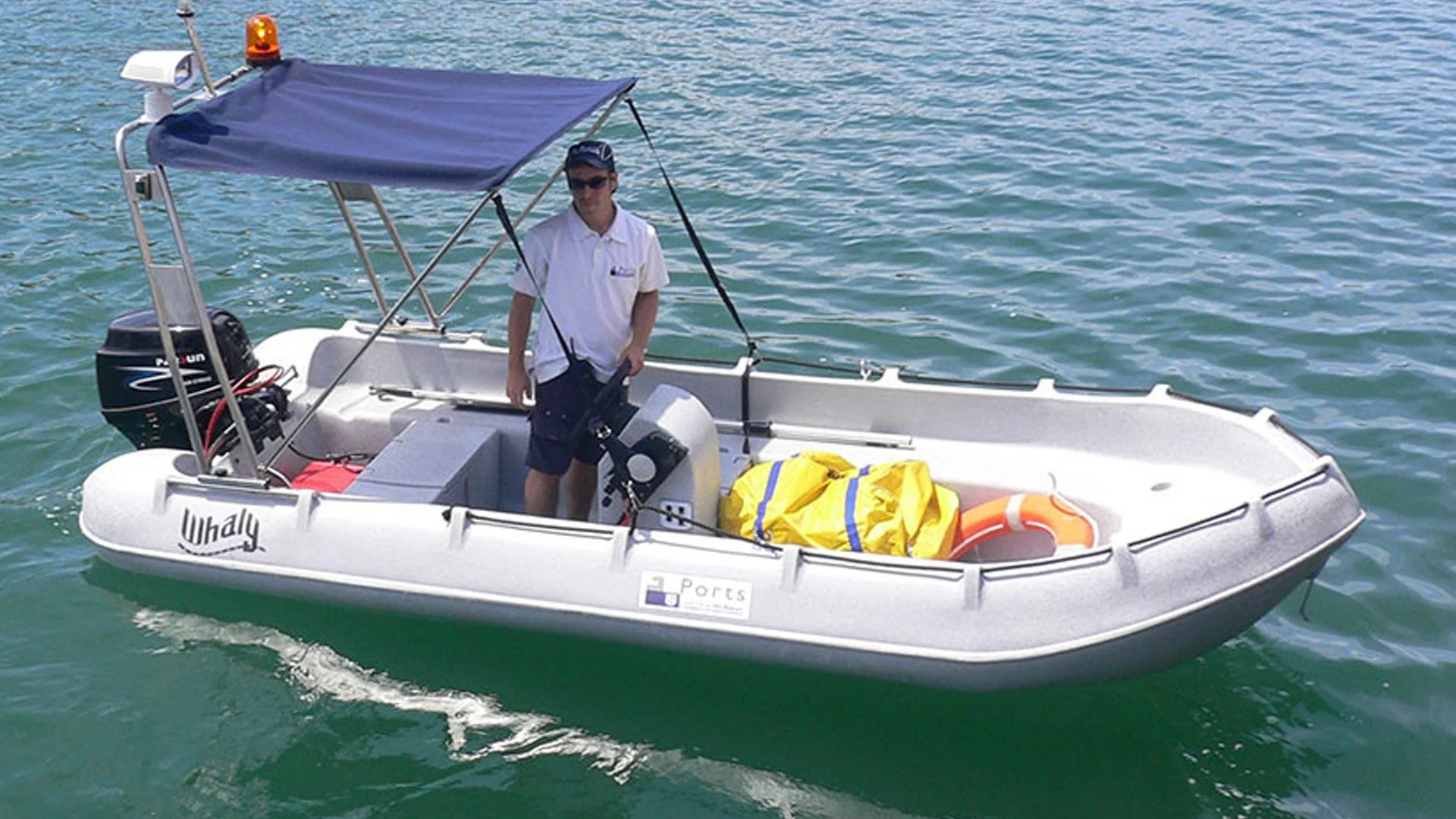 A man on a small white boat with a blue canopy, out on the water, with a yellow life jacket and orange life ring visible.