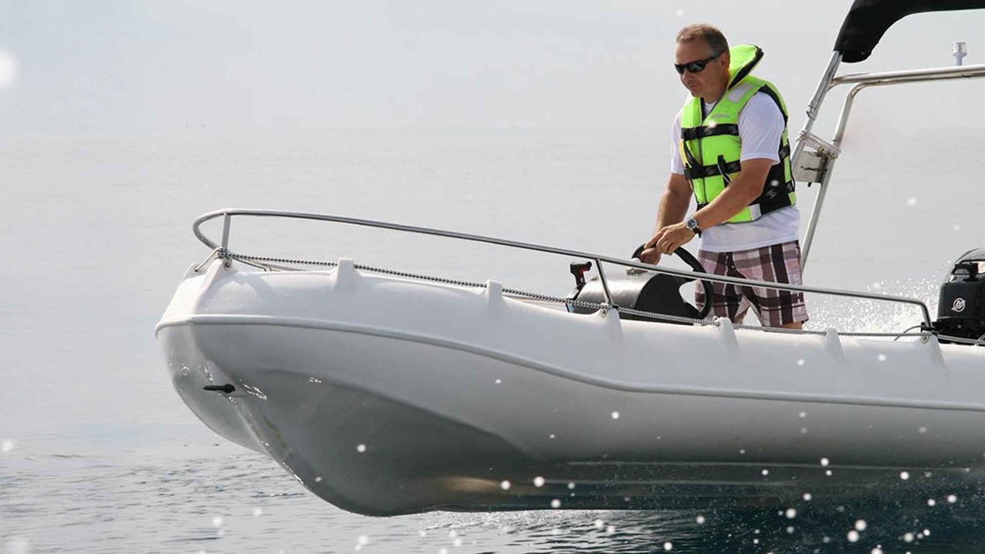 Man wearing sunglasses and a yellow life vest, standing on a small boat, steering with a wheel, dressed in a white T-shirt and plaid shorts, on a body of water.