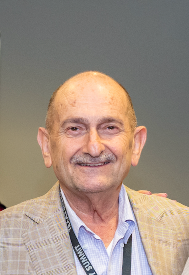 Portrait of an elderly man smiling, wearing a beige plaid blazer and a light blue collared shirt, with a lanyard around his neck.