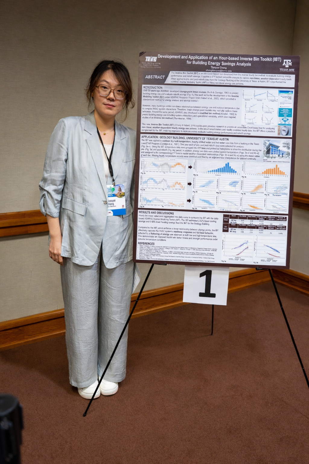 A woman with glasses and a light gray suit stands next to a large research poster at a conference, with her left arm resting at her side and a name badge hanging around her neck. The poster includes graphs and text related to energy savings research.
