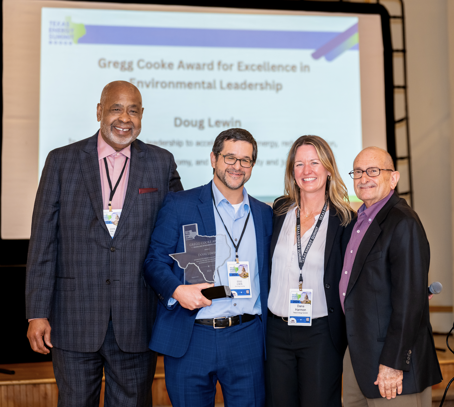 Four people standing together in front of a large screen at an award ceremony. The person second from the left is holding a trophy shaped like the state of Texas. The screen displays text about the Gregg Cooke Award for Excellence in Environmental Le