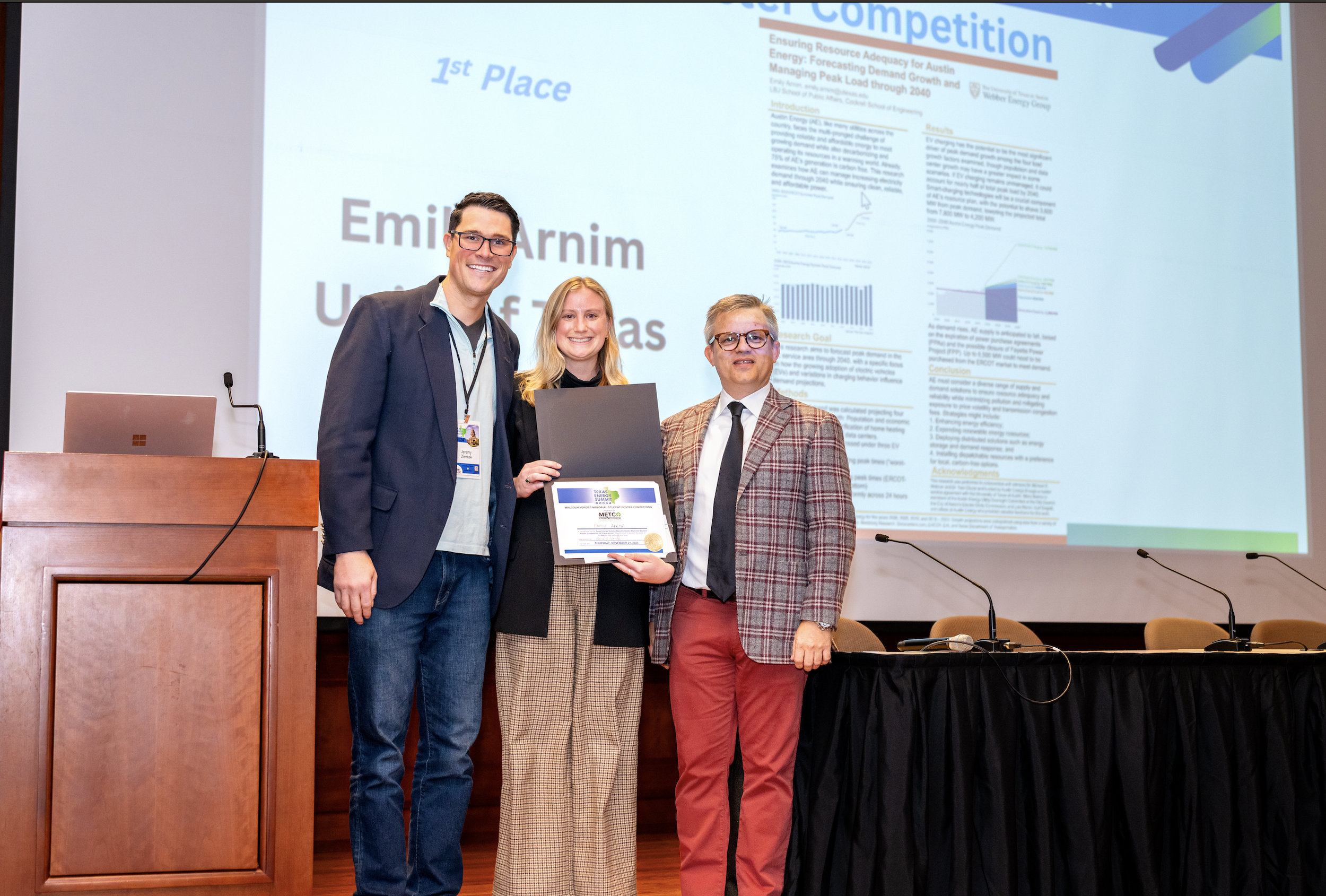 Three people standing in front of a presentation screen at an award ceremony, with the woman in the middle holding a certificate and medal. The presentation slide behind them shows '1st Place' and 'Emilio Arnim University of Texas'. A wooden podium a
