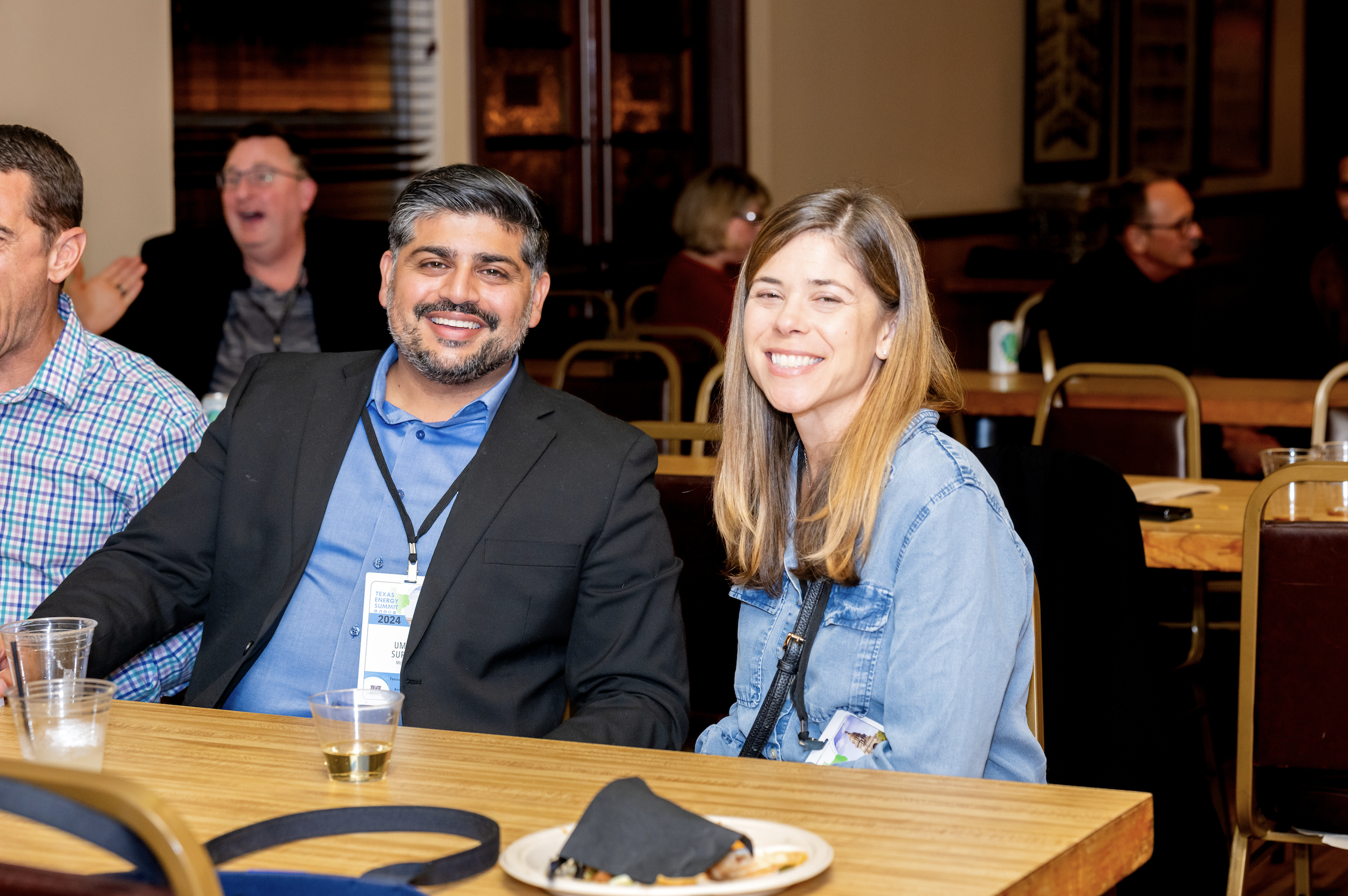 Two people sitting at a table, smiling and looking at the camera, with others in the background. The man has dark hair and a beard, wearing a black blazer and blue shirt. The woman has long light brown hair, wearing a denim jacket. They are at a soci