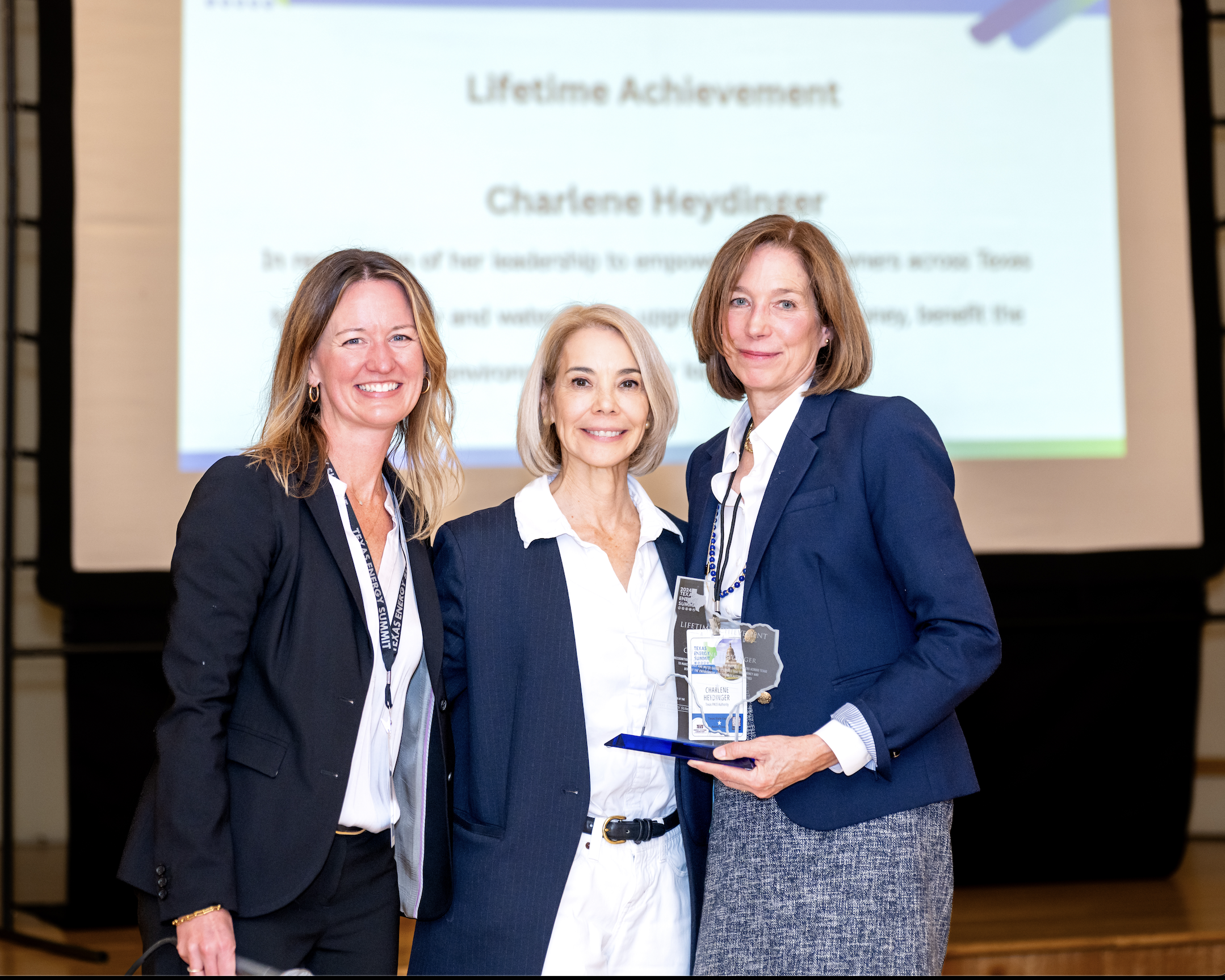Three women in business attire standing together on stage, smiling, with a presentation slide behind them that reads "Lifetime Achievement" and "Charlene Heydinger".