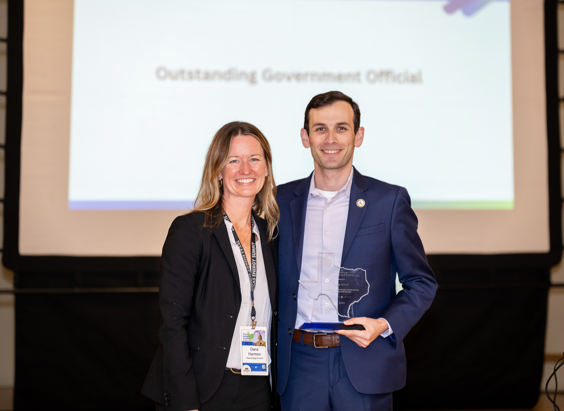 Two people, a woman and a man, standing together in front of a screen. The woman has blonde hair, is wearing a black blazer, and a conference badge. The man has dark hair, is in a blue suit, and is holding a clear award that has a Texas outline and t