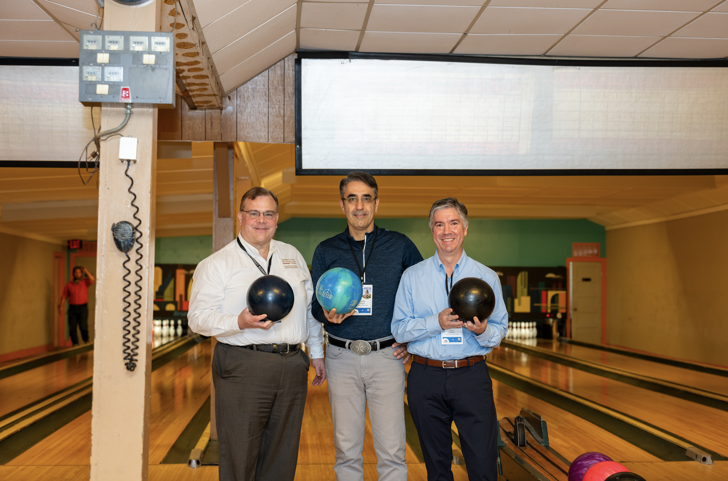 Three men standing in a bowling alley holding bowling balls, with a woman in a red shirt in the background near the lanes.
