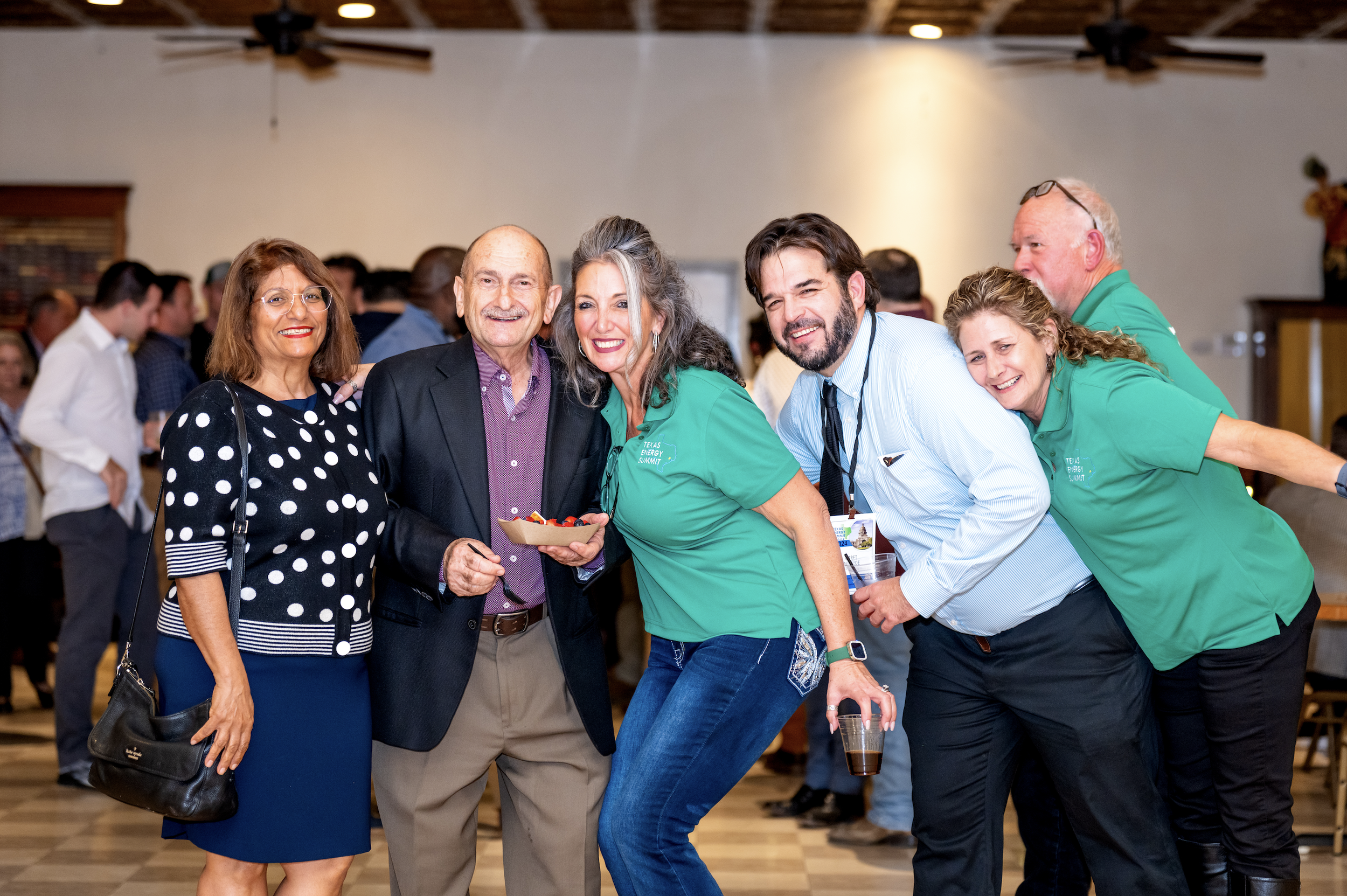 Group of six people smiling and posing together at an indoor event, with some holding drinks, in a festive atmosphere.