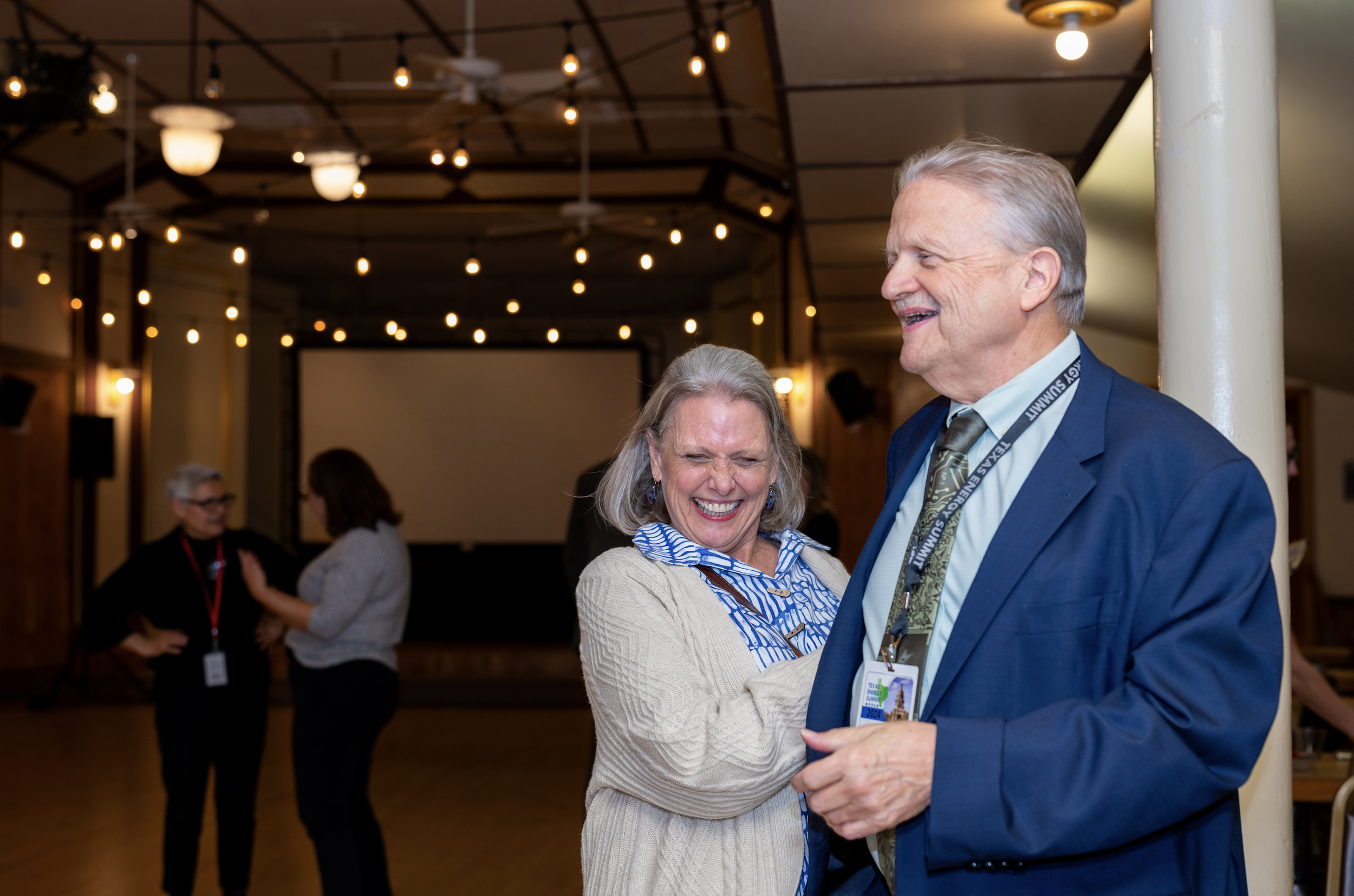 An elderly couple dancing and smiling at a social event in a decorated indoor venue with string lights and a large screen in the background.