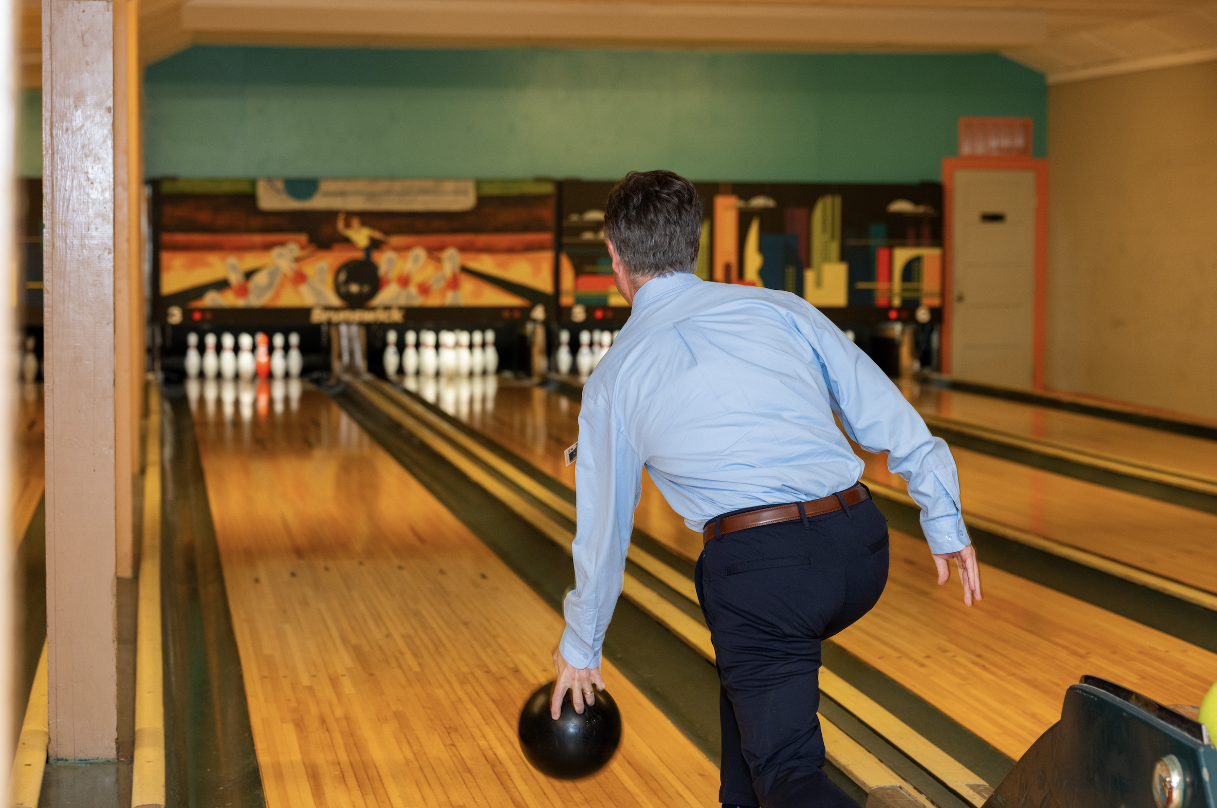 A man in a light blue shirt and dark pants prepares to roll a black bowling ball down a lane at a bowling alley, with pins set up at the end of the lane.