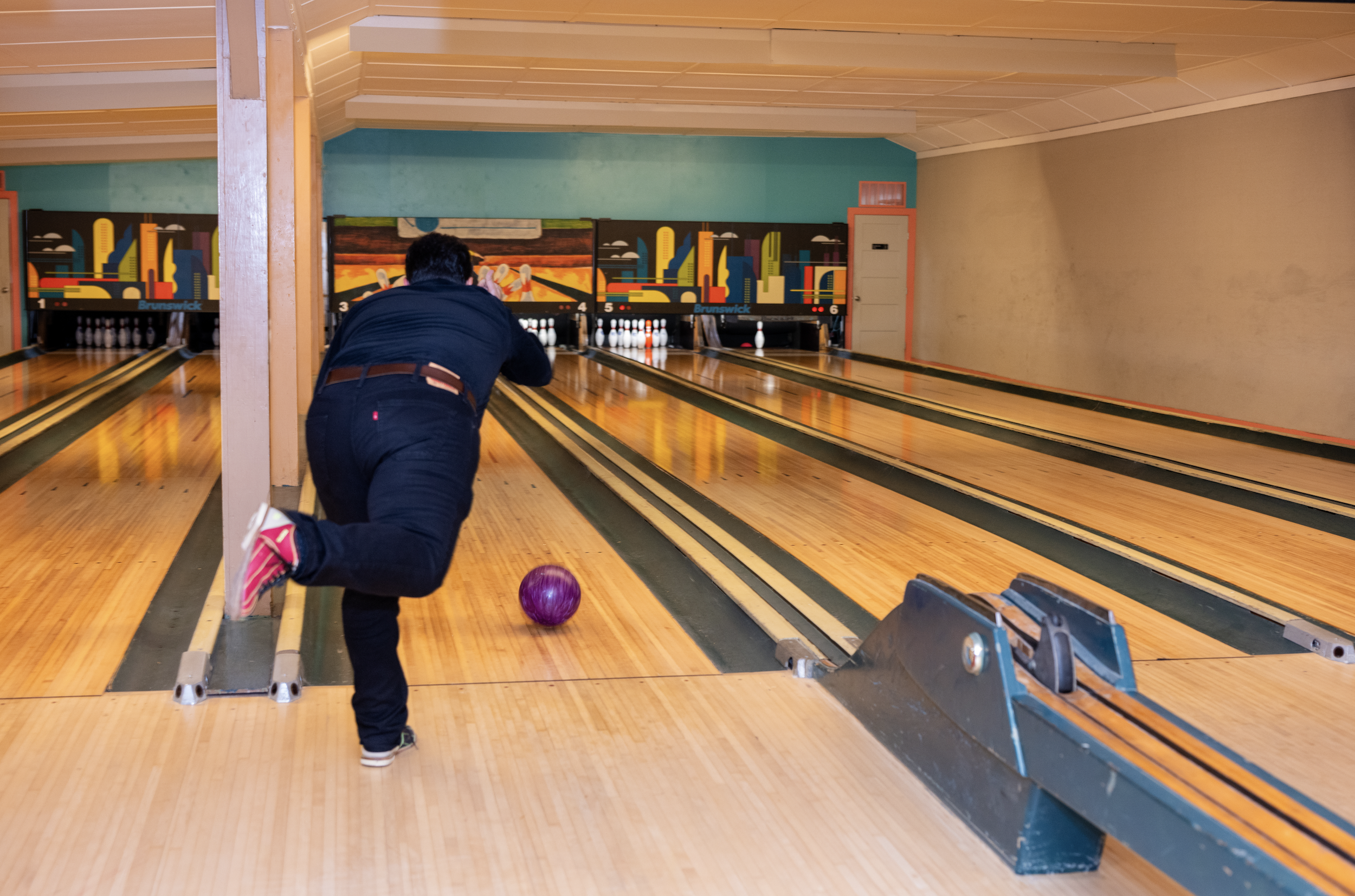 A man bowling in an indoor bowling alley with a purple bowling ball rolling down the lane towards the pins.