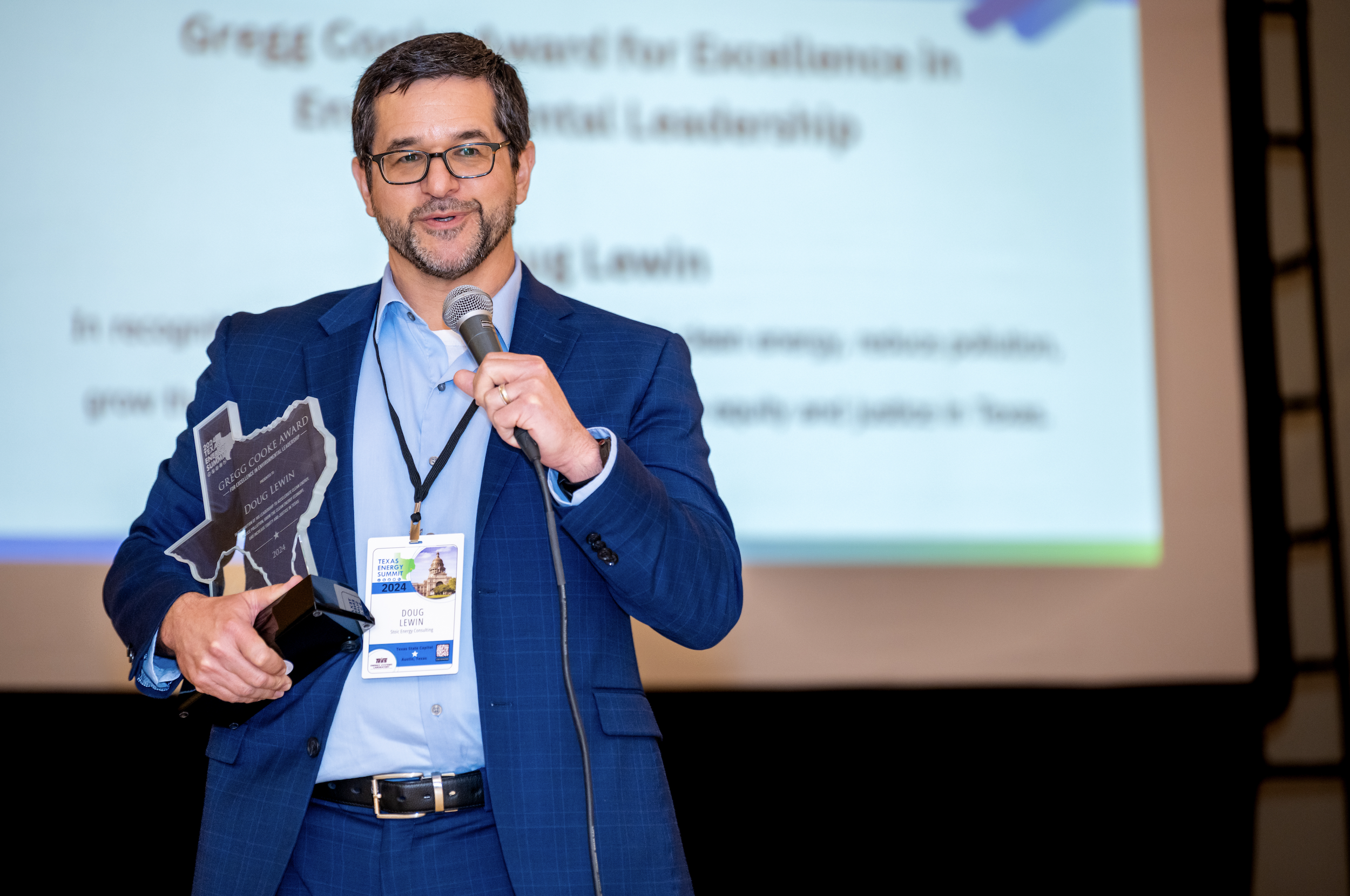 A man wearing a blue suit and glasses holding a microphone and an award shaped like Texas, with a scene of the Texas Capitol building on his conference badge. He is standing in front of a presentation slide at the Texas Energy Summit 2024.